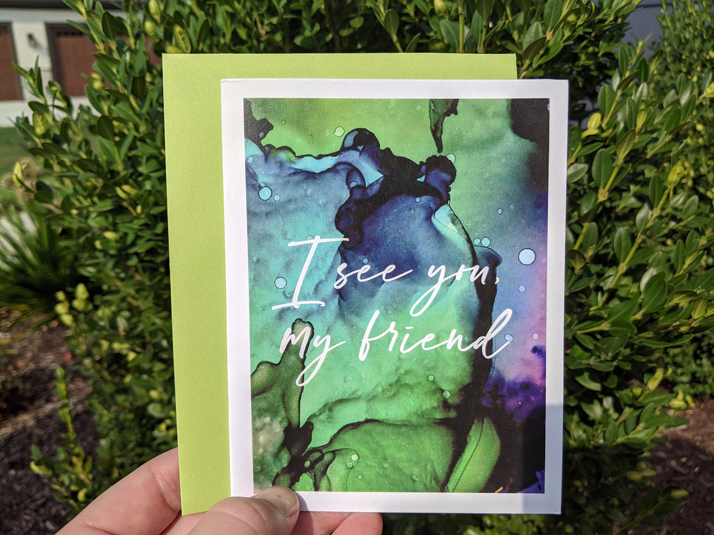 Hand holding a greeting card and colorful envelope in front of evergreens outdoors.
