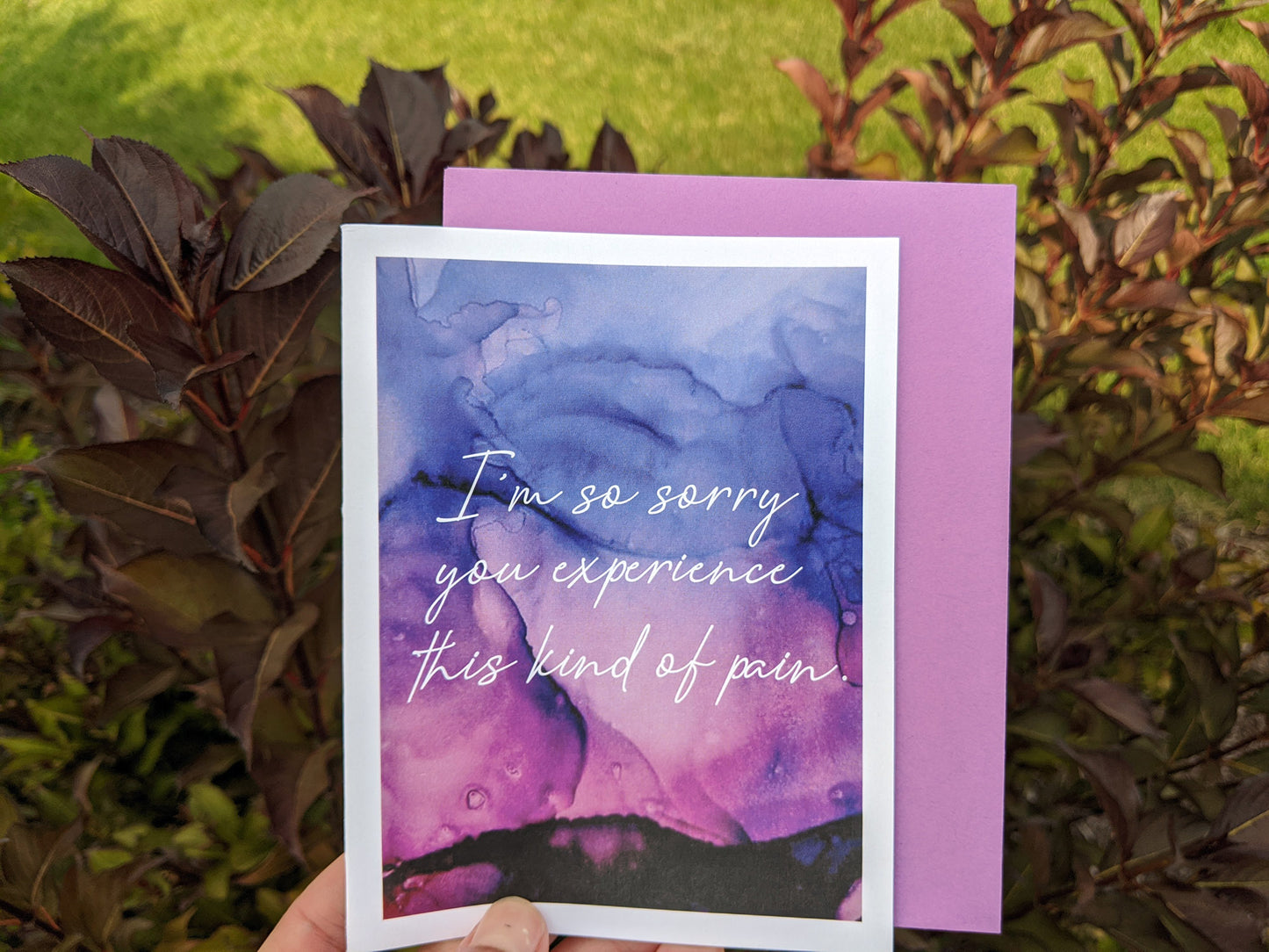 Hand holding a greeting card and kraft envelope in front of shrubs outdoors.