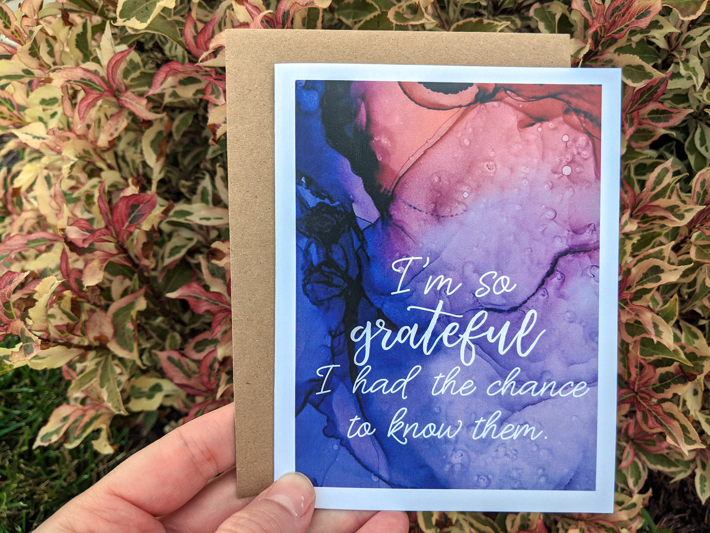 Hand holding a greeting card and kraft envelope in front of shrubs outdoors.