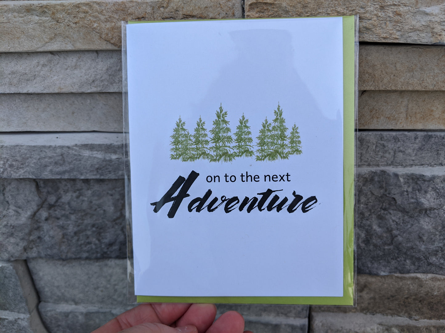 Hand holding a greeting card and colorful envelope in front of stones outdoors.