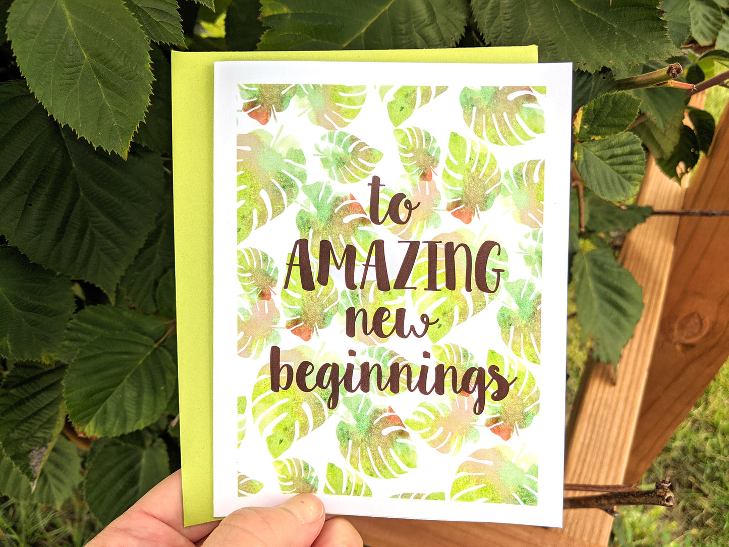 Hand holding a greeting card and colorful envelope in front of shrubs outdoors.