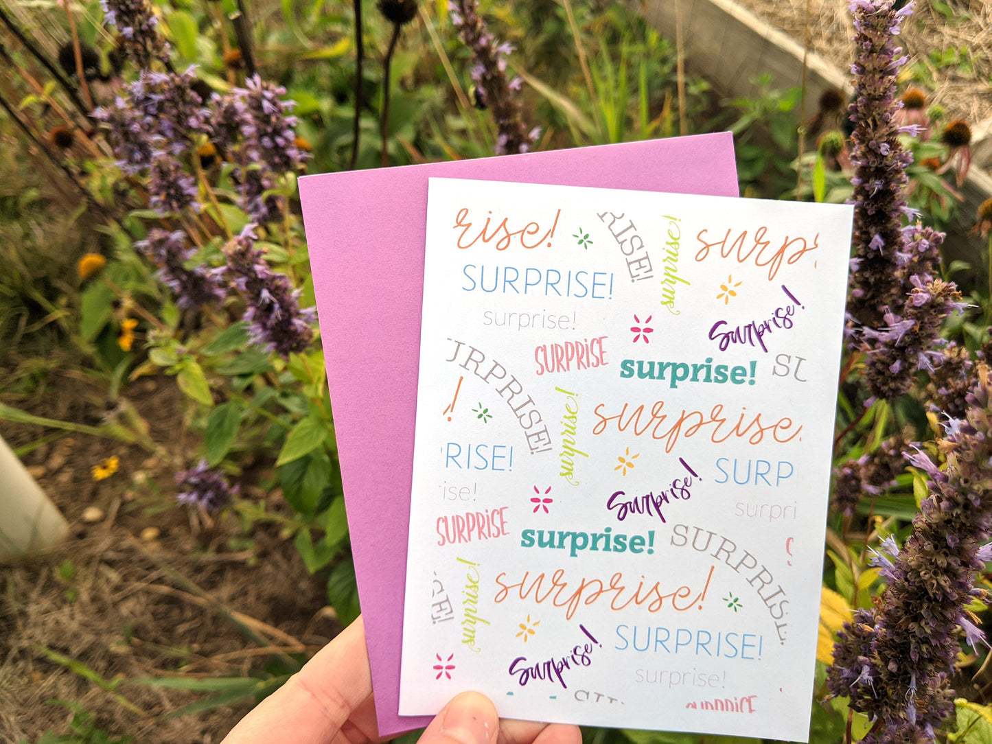 Hand holding a greeting card and colorful envelope in front of shrubs outdoors.
