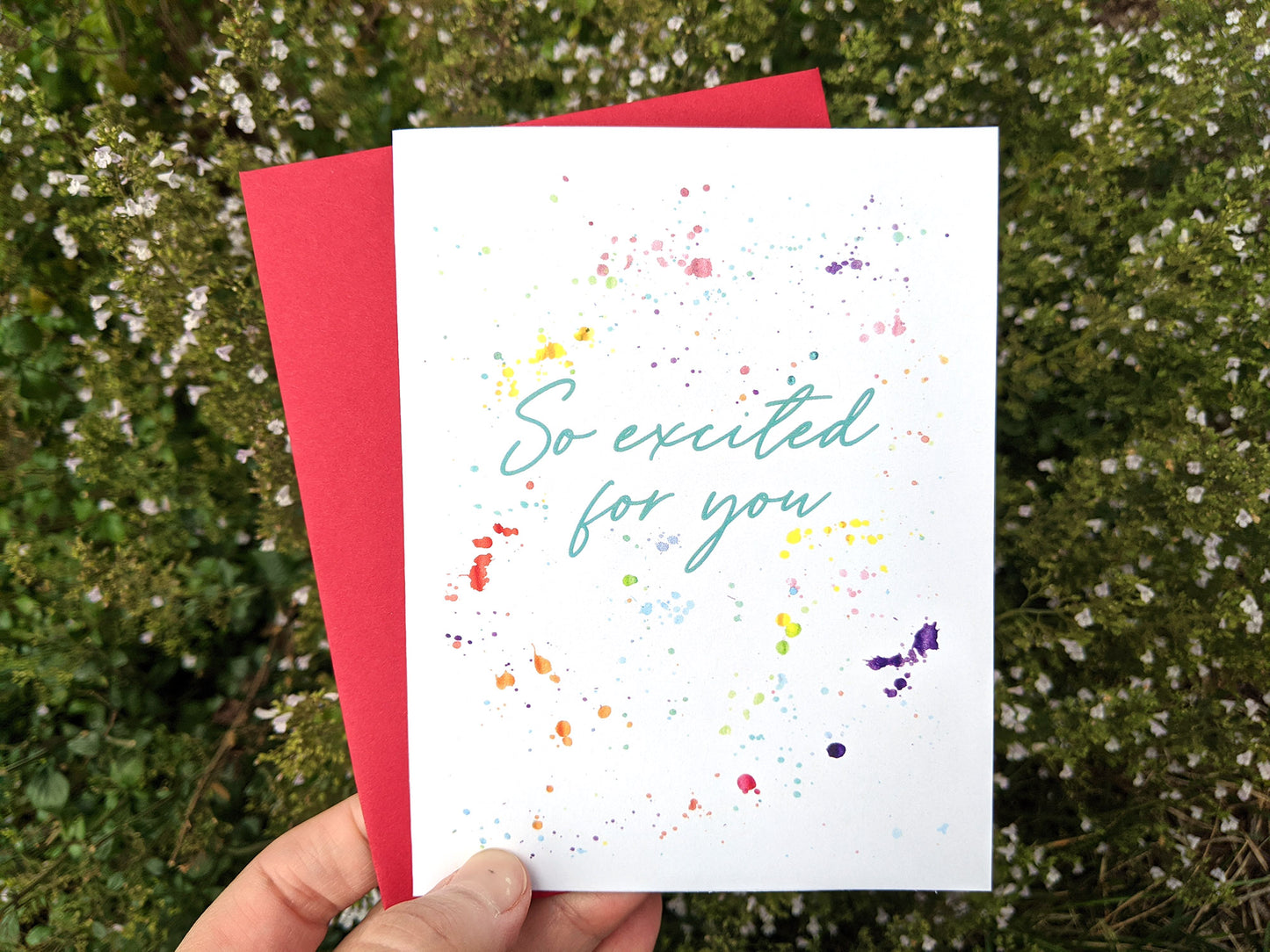Hand holding a greeting card and colorful envelope in front of shrubs outdoors.