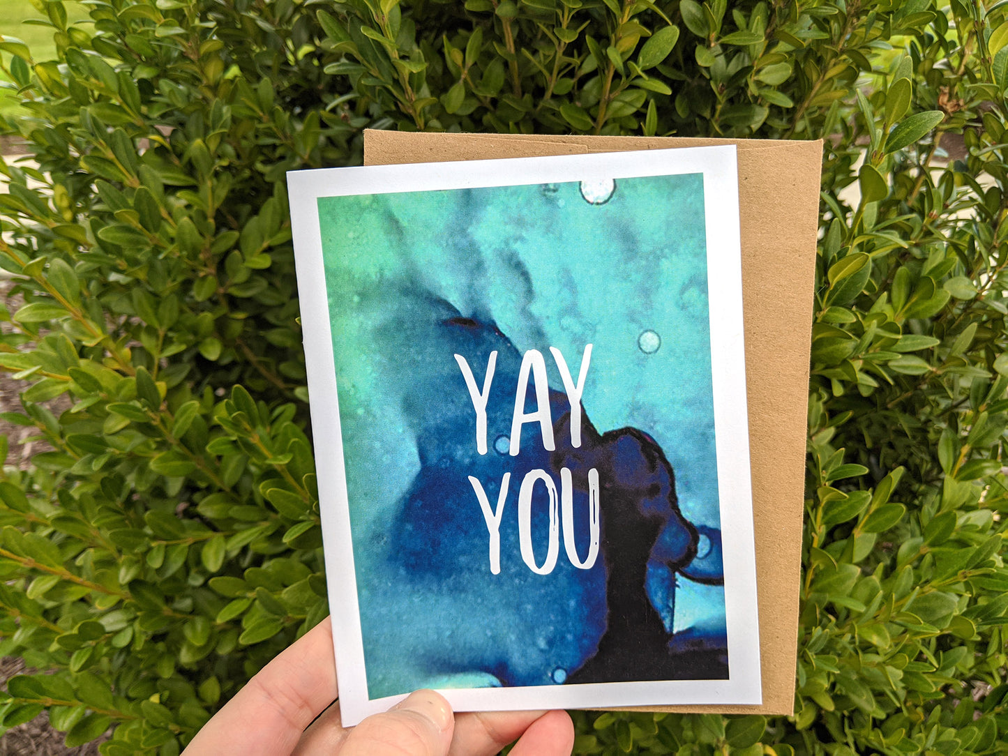 Hand holding a greeting card and kraft envelope in front of shrubs outdoors.