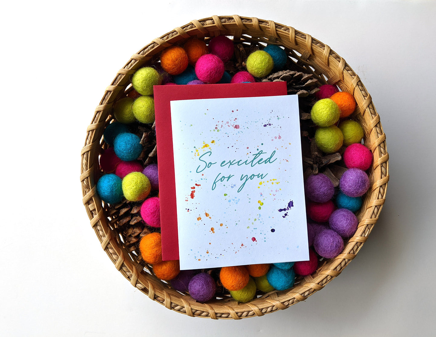 Greeting card laying in a woven basket filled with colorful felt balls and pinecones.