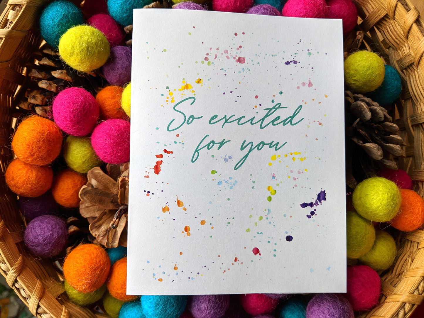 Greeting card laying in a woven basket filled with colorful felt balls and pinecones.