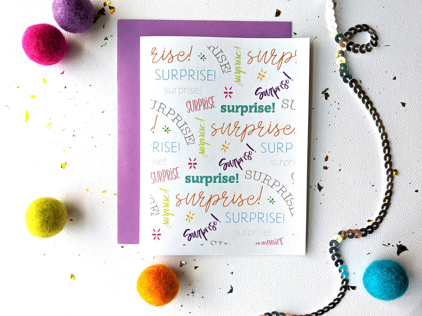 Greeting card with a colorful envelope laying on a white surface with colorful felt balls and a string of sequins around it.
