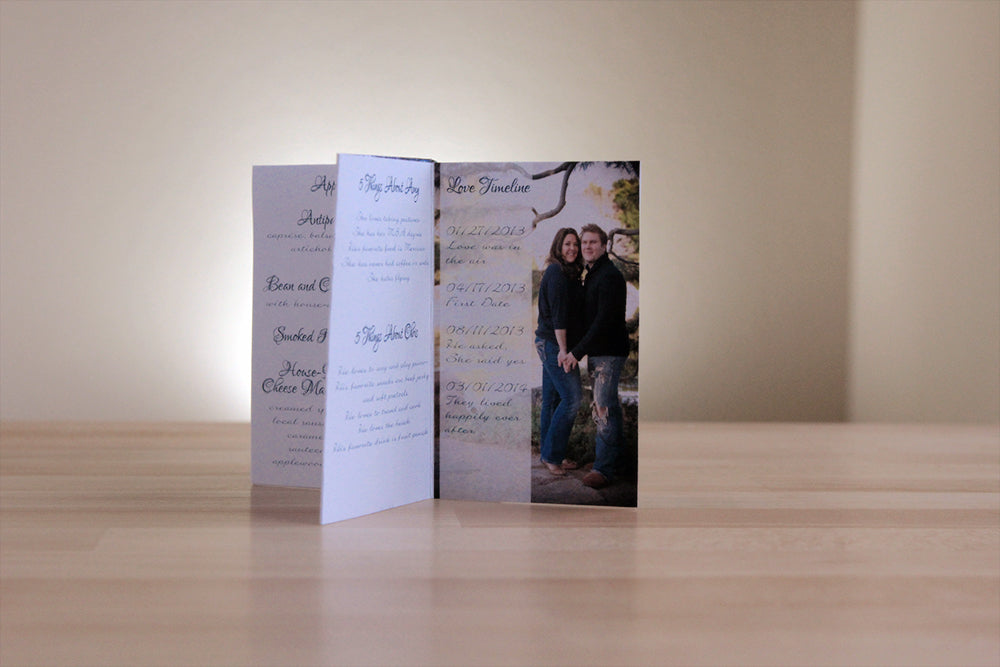 Centerpiece sitting on a wooden tabletop with panels showing an engagement photo overlaid with their relationship story timeline, trivia facts about the couple and part of a menu.
