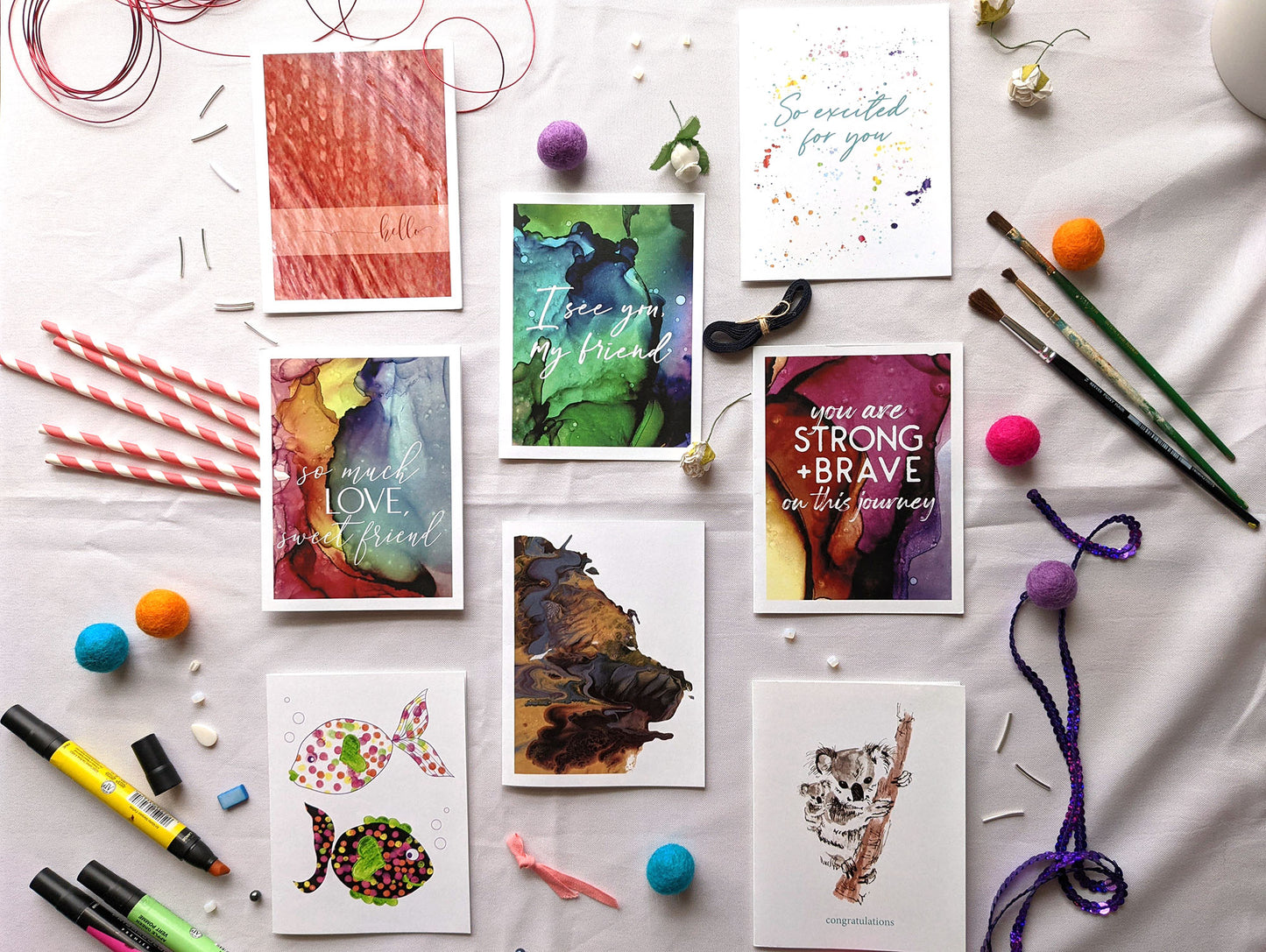 Flatlay of 8 greeting cards in three columns surrounded by markers, paper straws, beads, paintbrushes, colorful felt balls, and sequins.