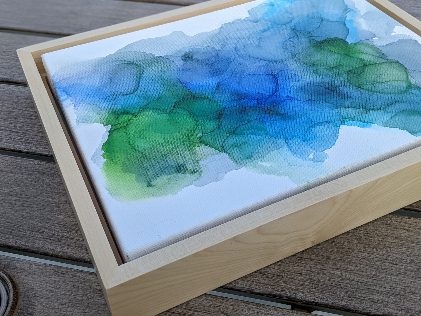 Photo angled from above of the canvas in a light maple float frame laying on a slat patio table.