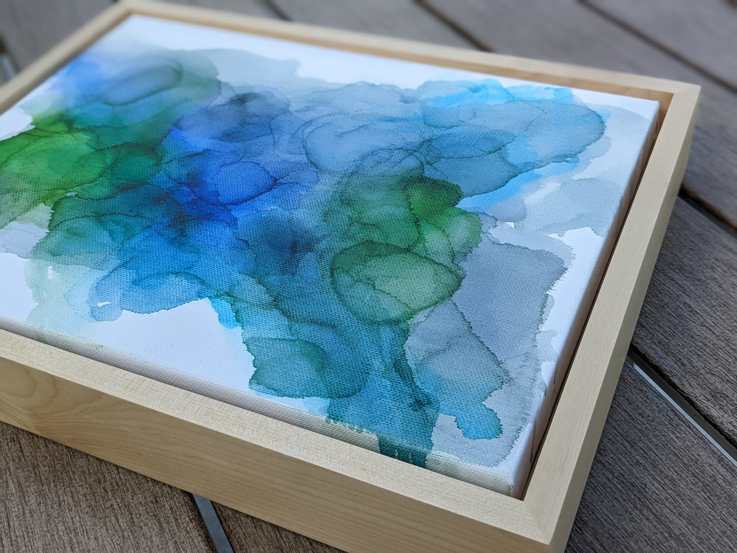 Photo angled from above of the canvas in a light maple float frame laying on a slat patio table.