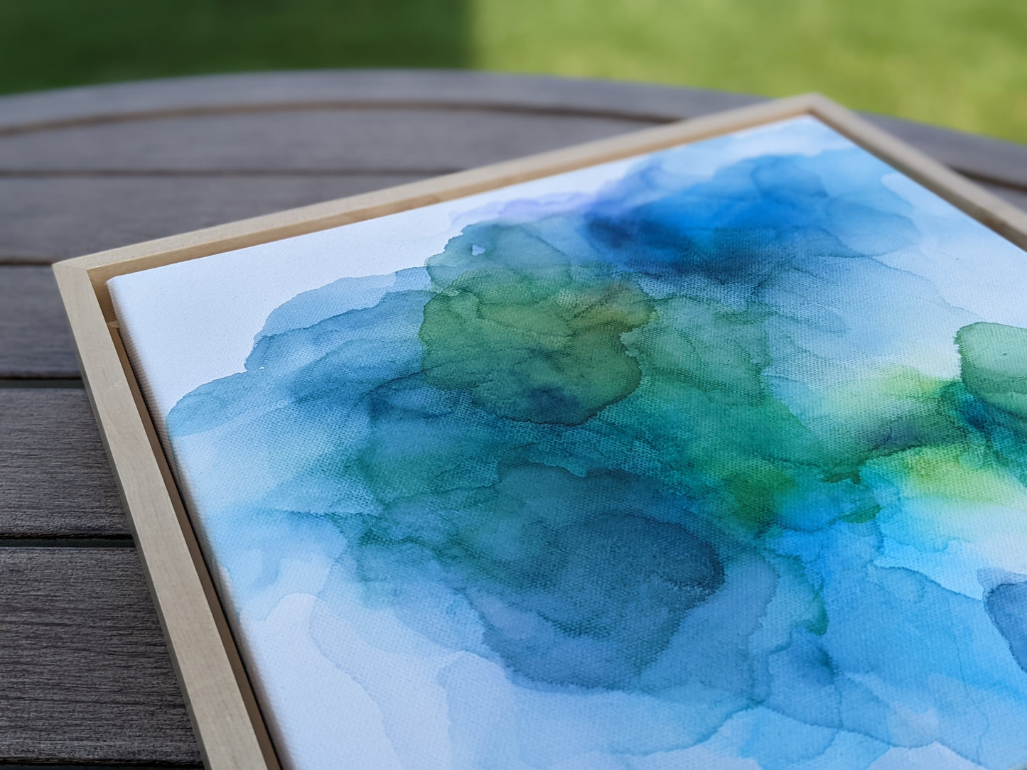 Photo angled from above of the canvas in a light maple float frame laying on a slat patio table.