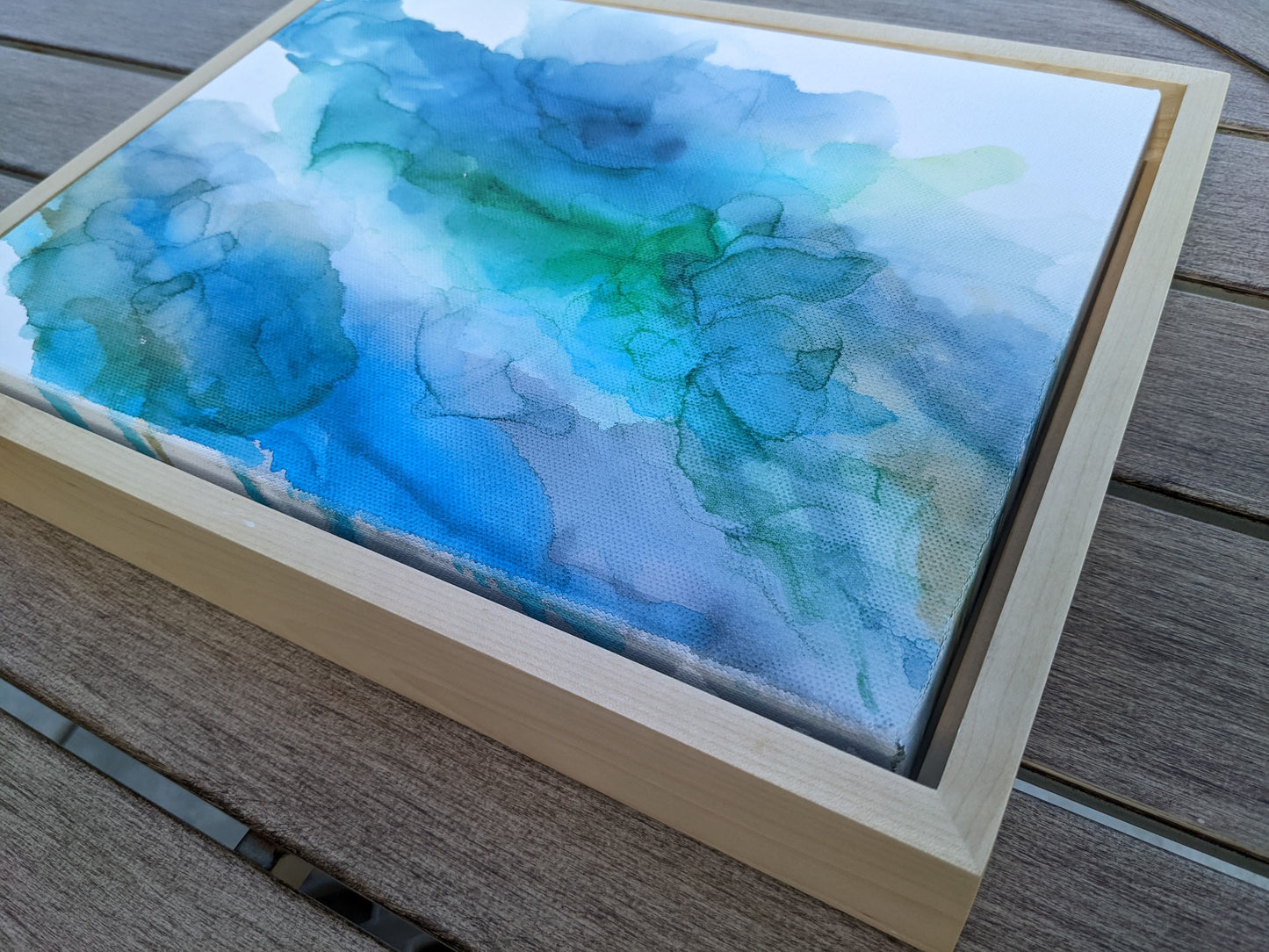 Photo angled from above of the canvas in a light maple float frame laying on a slat patio table.