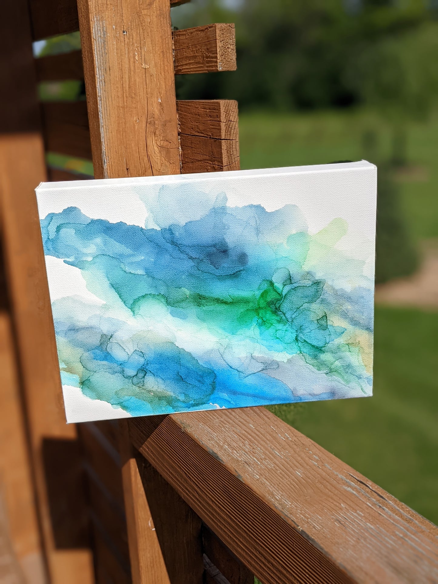 Canvas shown outdoors sitting on a wooden railing with greenery in the background.