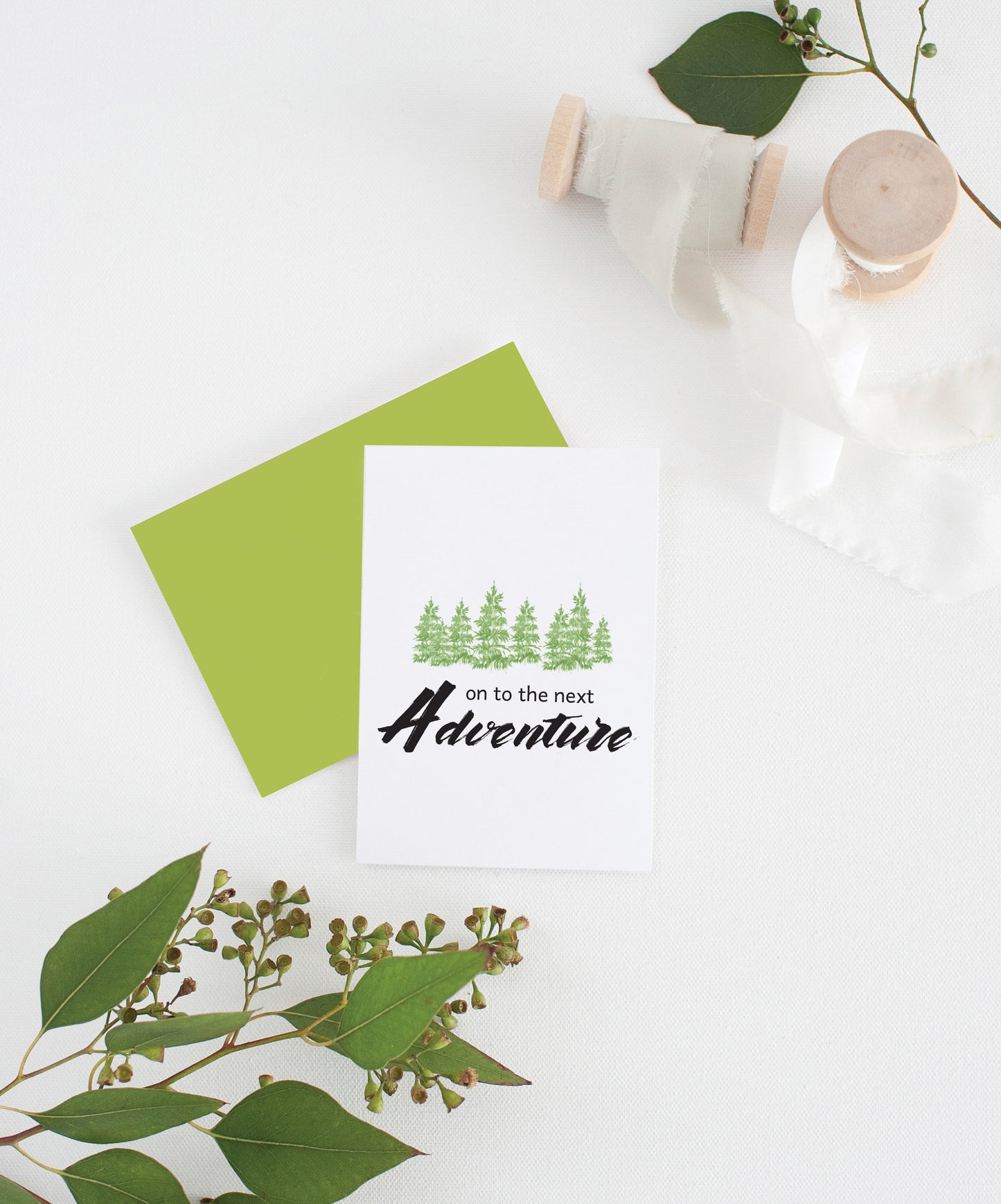 Greeting card with colorful envelope laying on a white surface with green leaves and cotton ribbon wound on wooden spools.