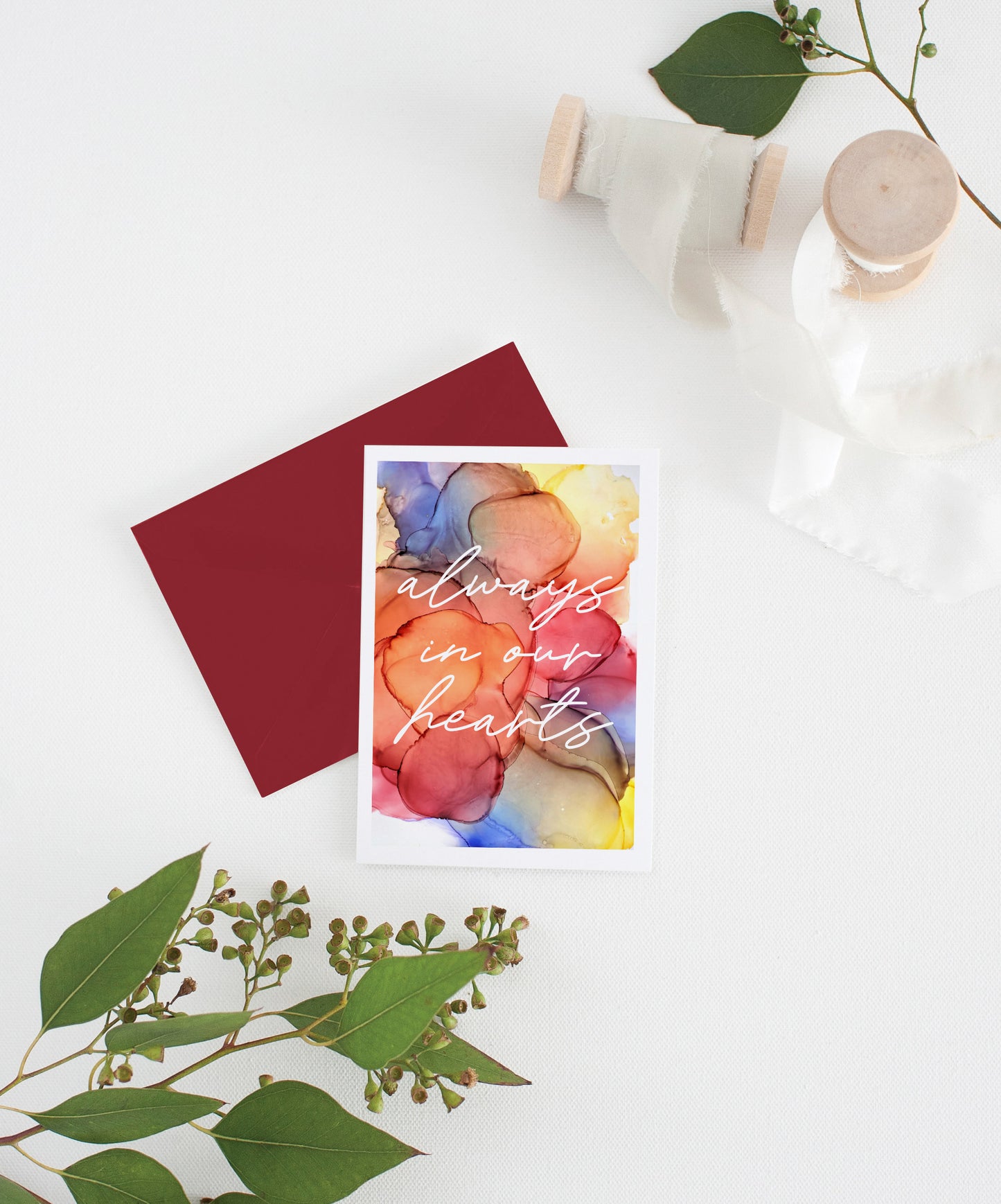 Greeting card with colorful envelope laying on a white surface with green leaves and cotton ribbon wound on wooden spools.