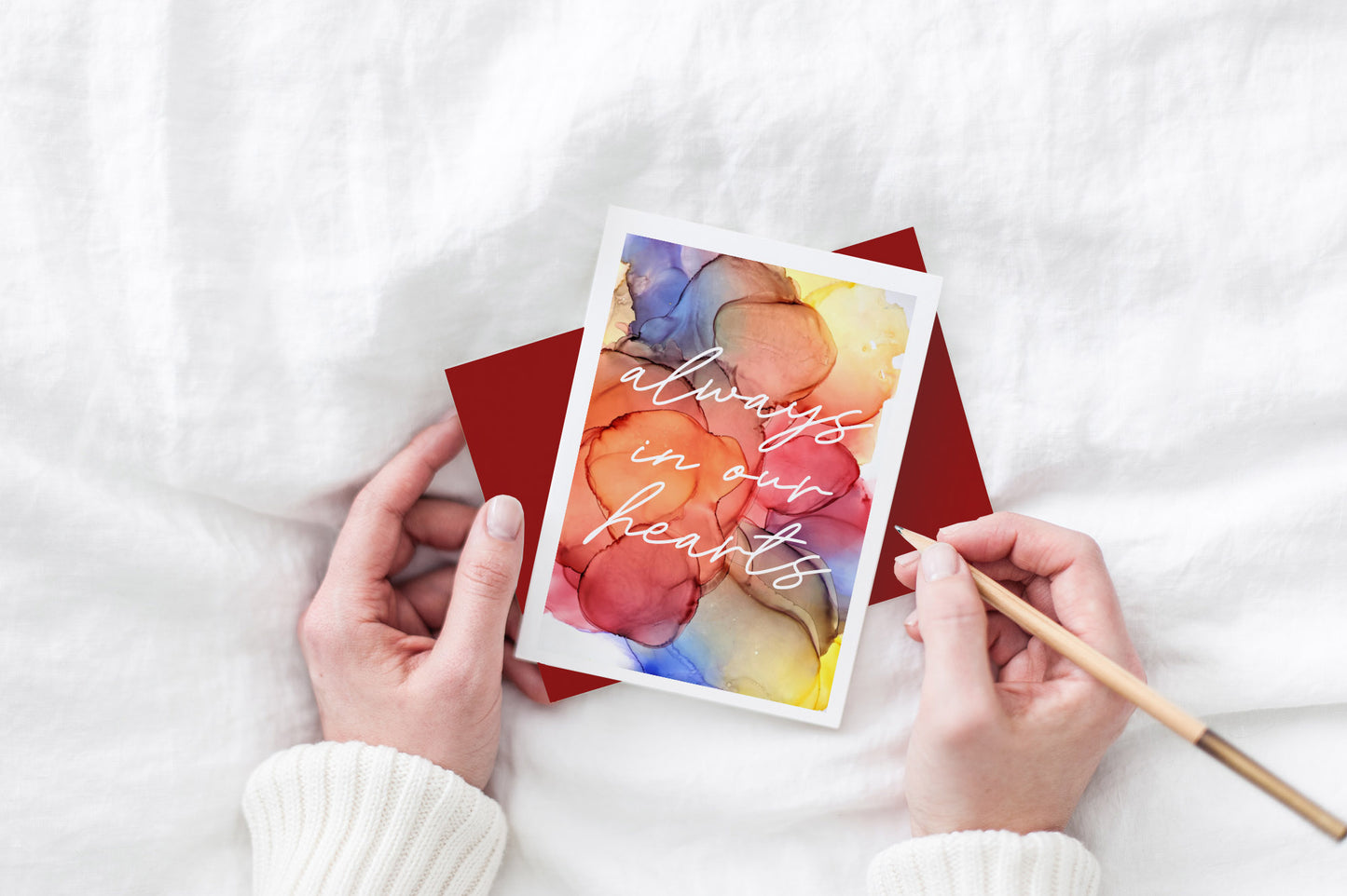 Greeting card and colorful envelope, seen from above, held by two hands along with a pencil, ready to write, against a white comforter.