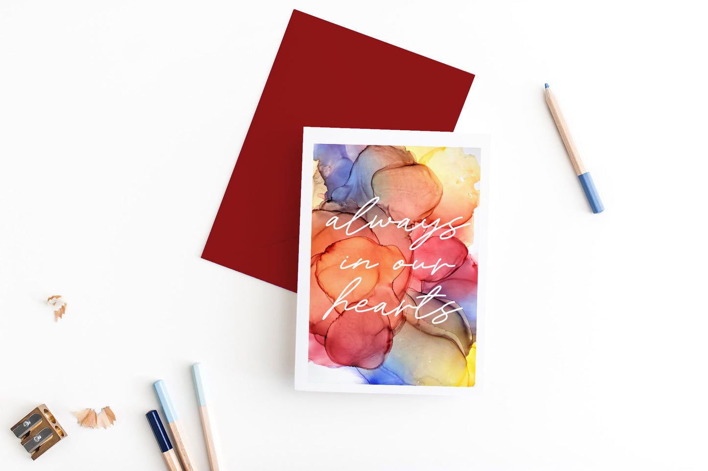 Greeting card, seen from above, with a colored, square flap envelope, pencils, pencil sharpener and shavings on a white background.