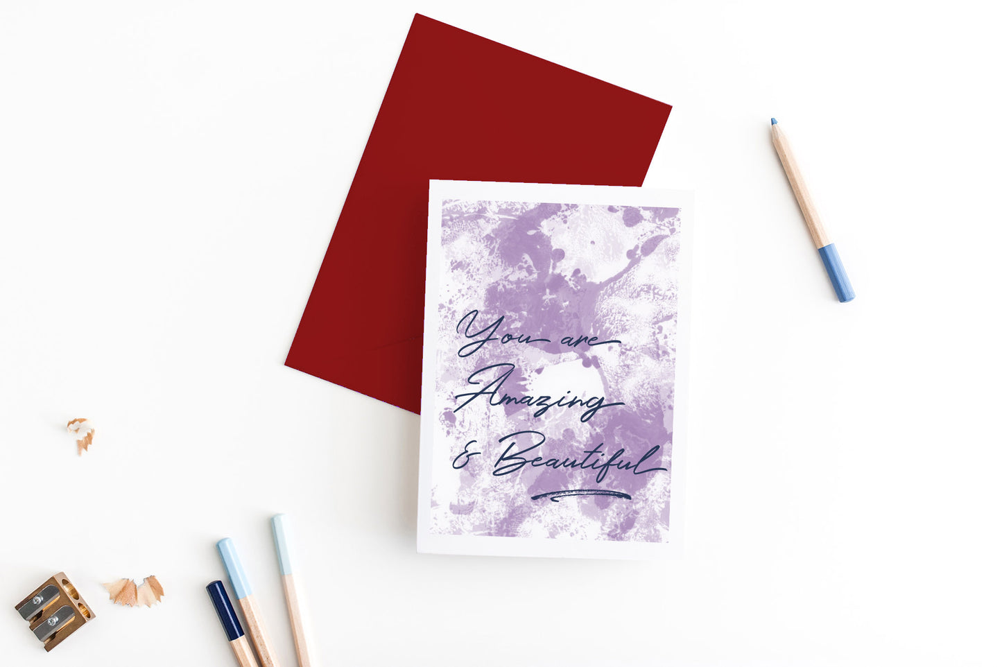 Greeting card, seen from above, with a colored, square flap envelope, pencils, pencil sharpener and shavings on a white background.