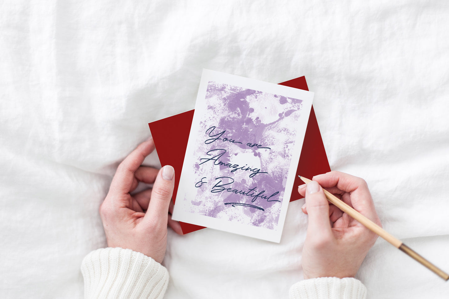 Greeting card and colorful envelope, seen from above, held by two hands along with a pencil, ready to write, against a white comforter.