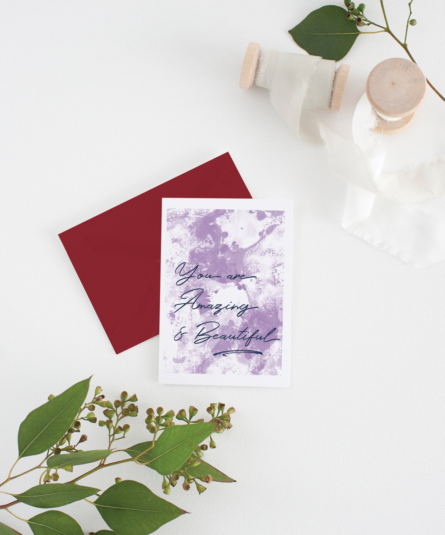 Greeting card with colorful envelope laying on a white surface with green leaves and cotton ribbon wound on wooden spools.