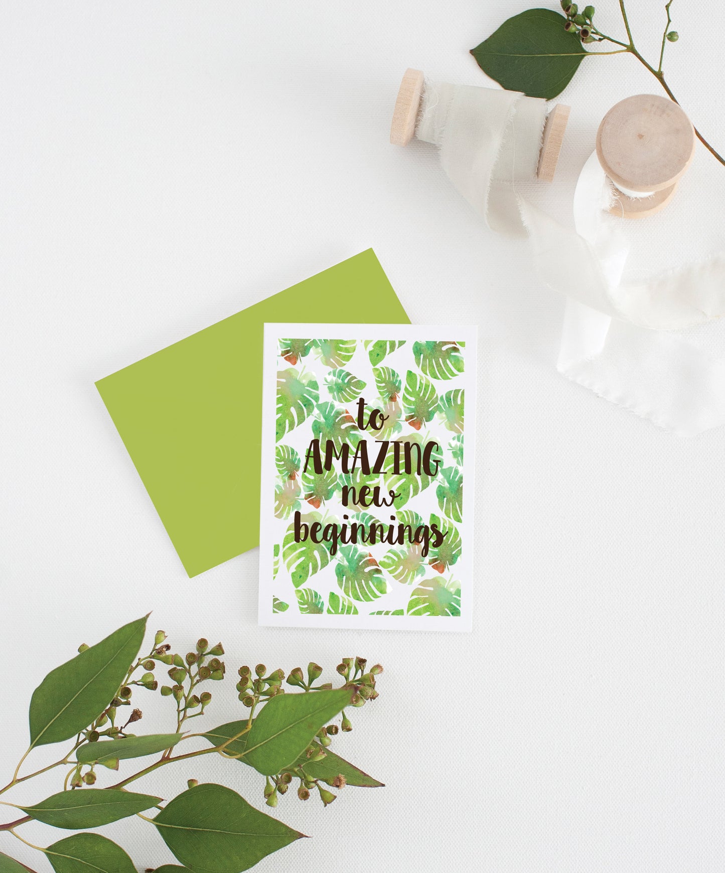 Greeting card with colorful envelope laying on a white surface with green leaves and cotton ribbon wound on wooden spools.