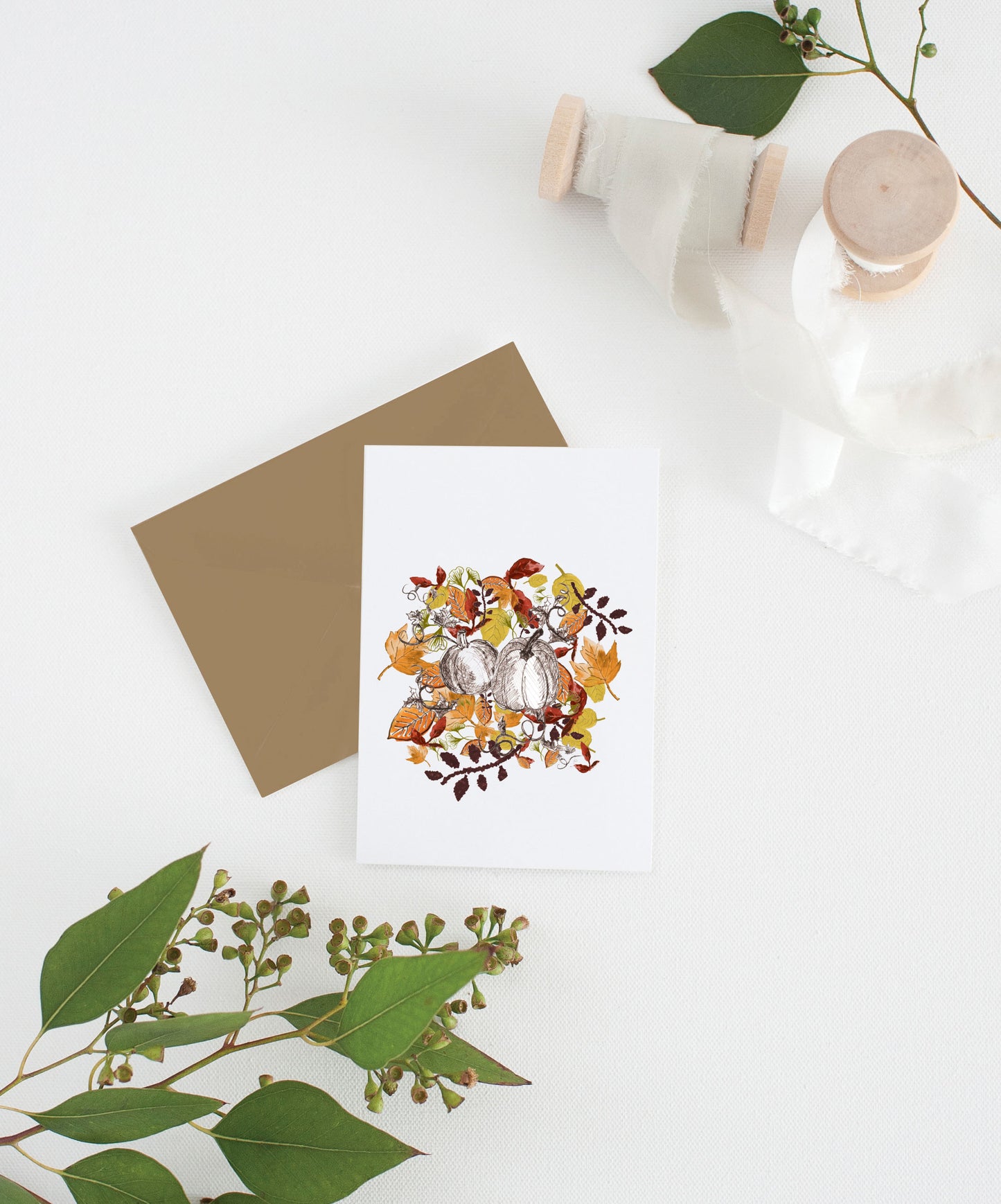 Greeting card with colorful envelope laying on a white surface with green leaves and cotton ribbon wound on wooden spools.