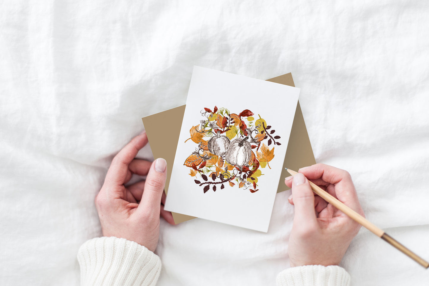Greeting card and colorful envelope, seen from above, held by two hands along with a pencil, ready to write, against a white comforter.
