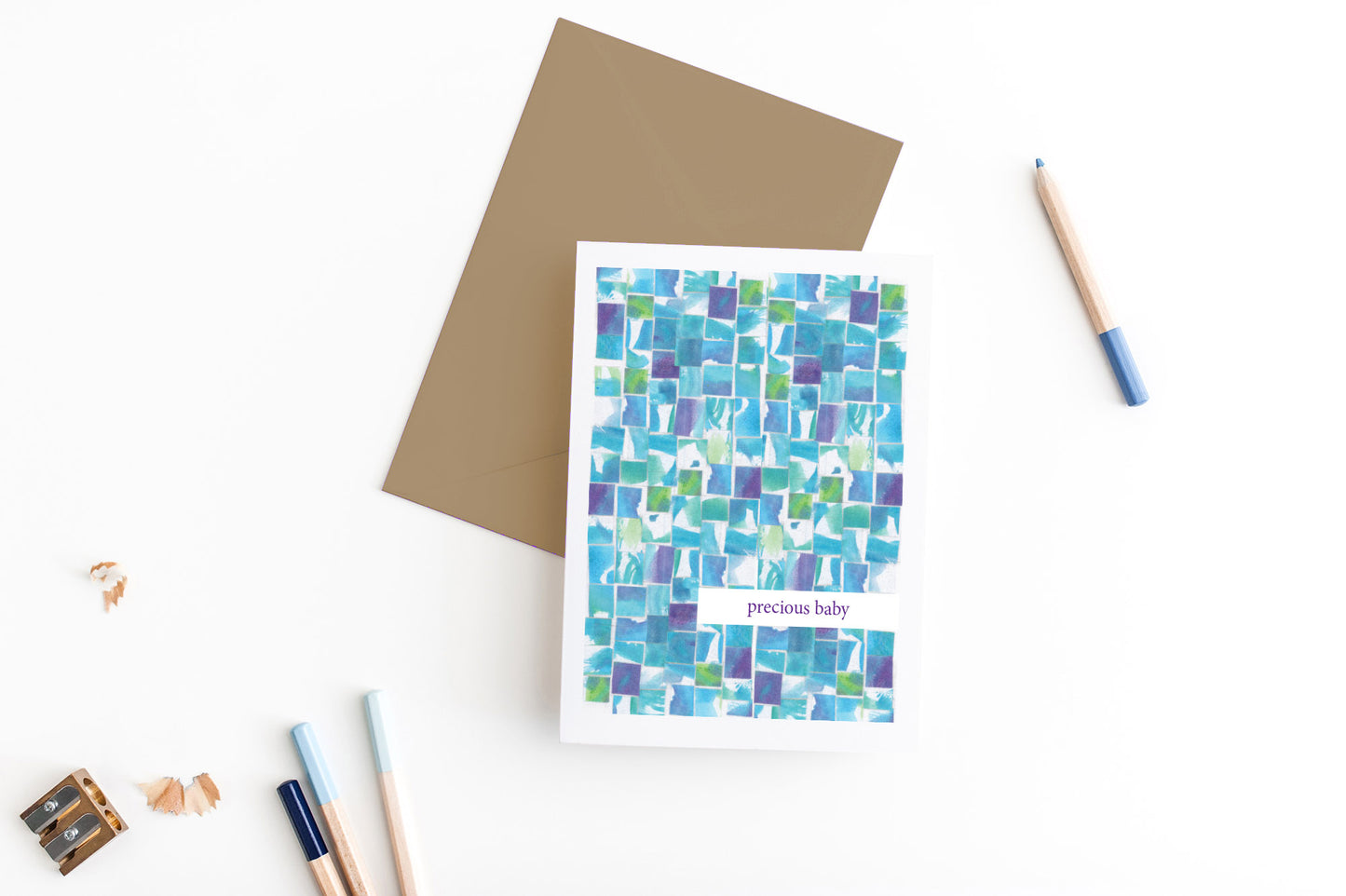 Greeting card, seen from above, with a kraft, square flap envelope, pencils, pencil sharpener and shavings on a white background.