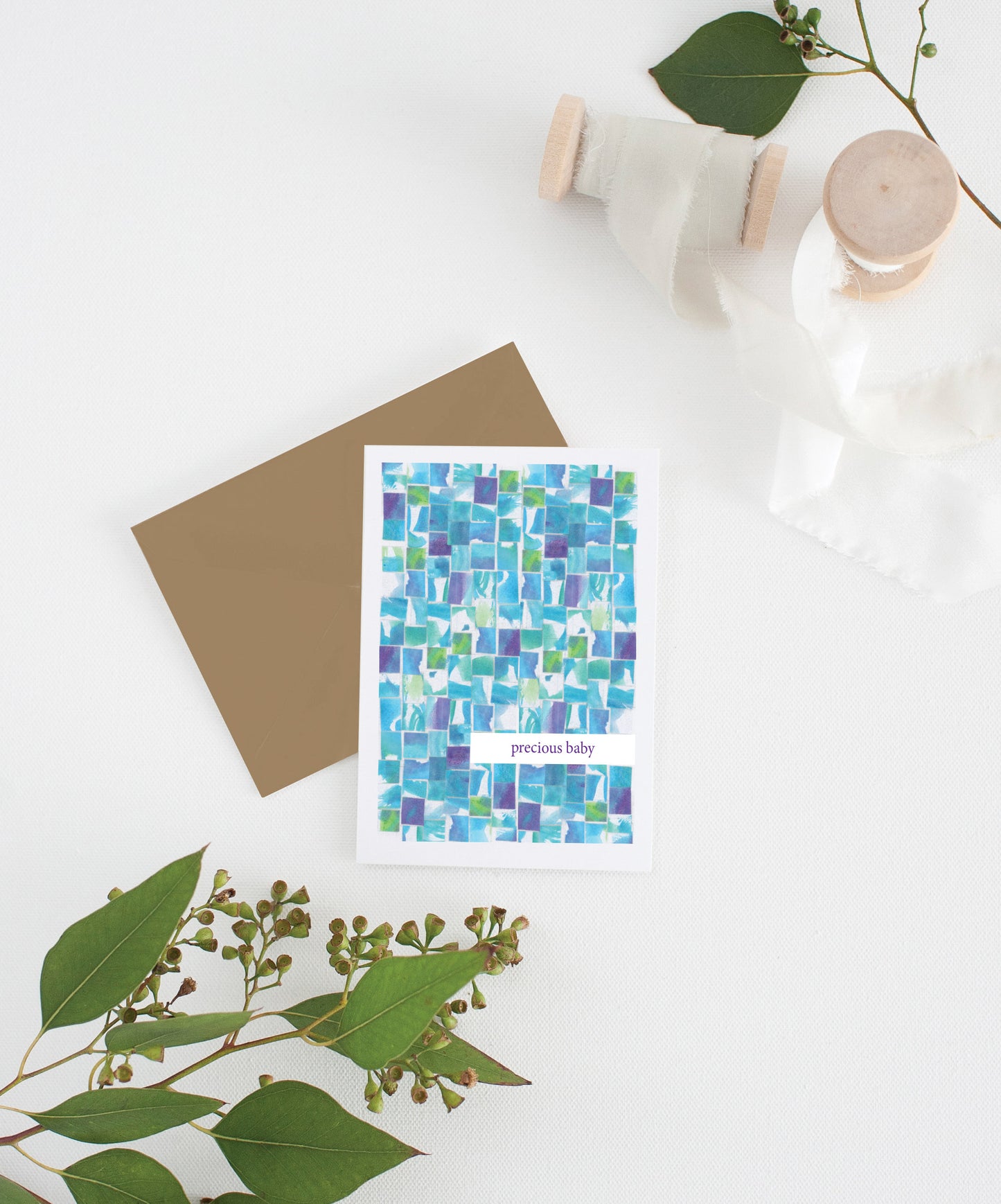 Greeting card with kraft envelope laying on a white surface with green leaves and cotton ribbon wound on wooden spools.