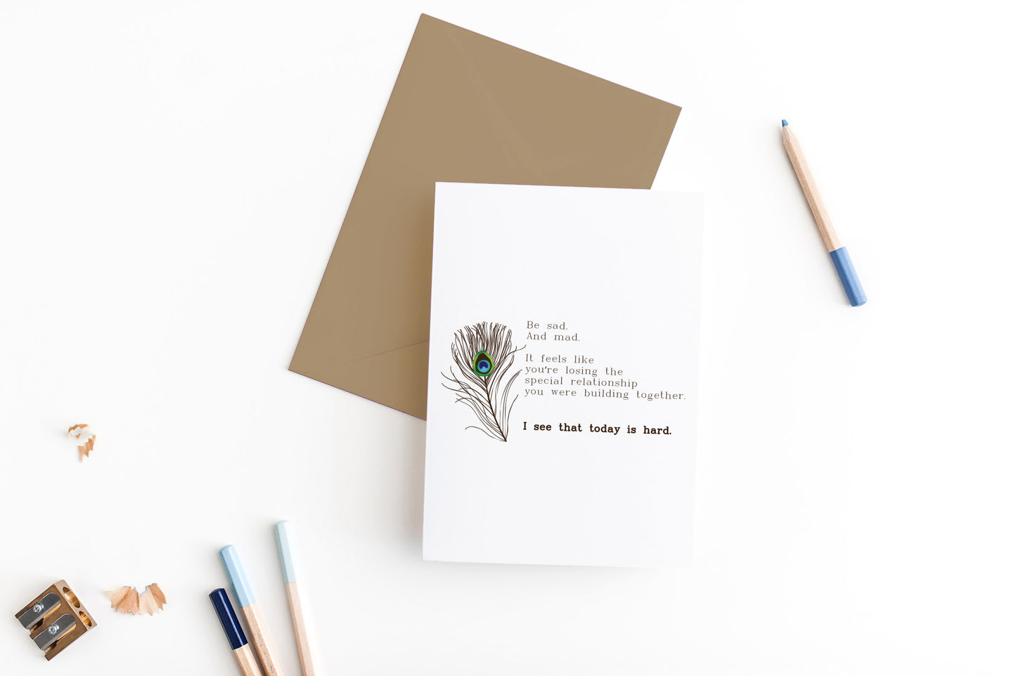Greeting card, seen from above, with a kraft, square flap envelope, pencils, pencil sharpener and shavings on a white background.
