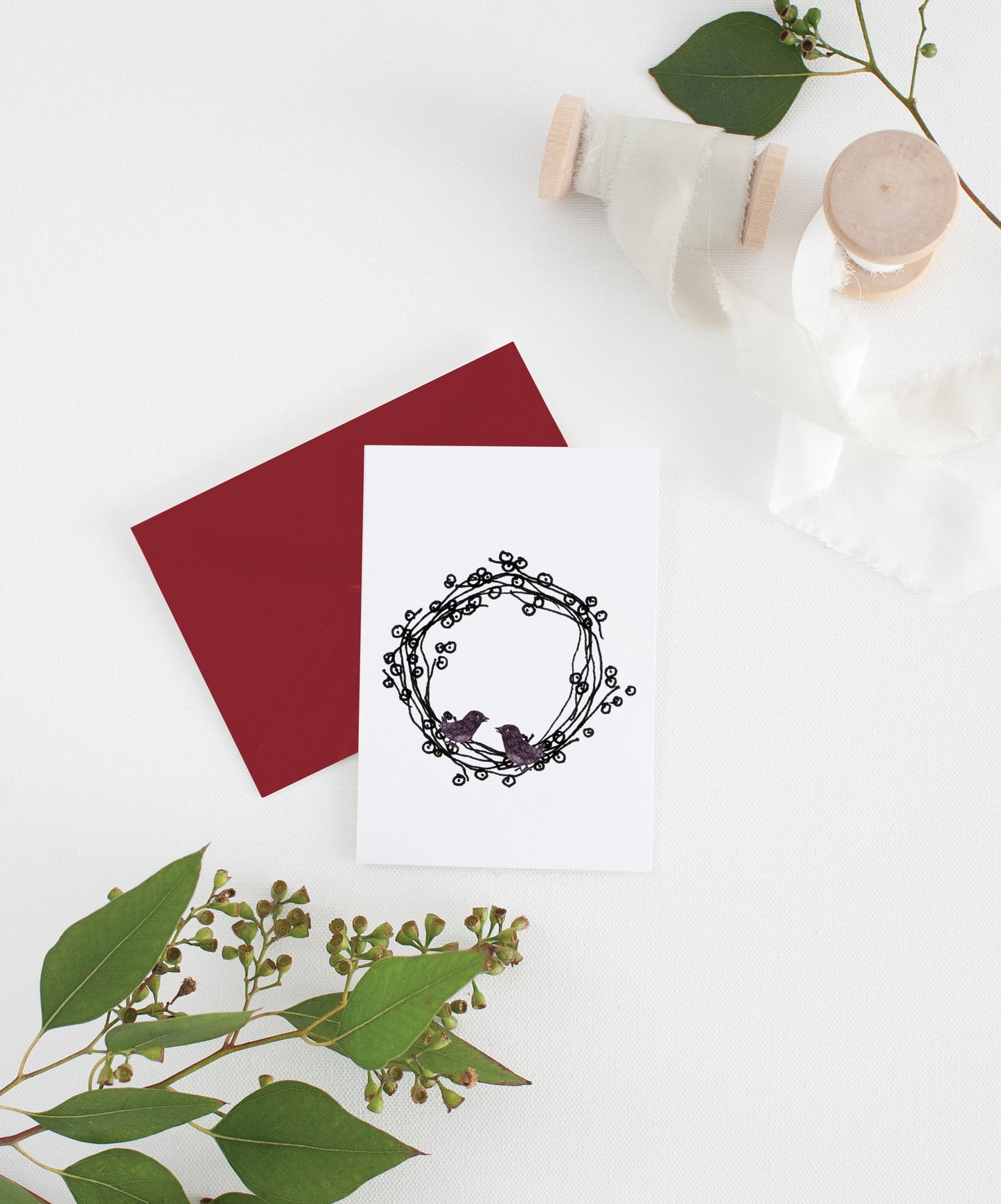 Greeting card with colorful envelope laying on a white surface with green leaves and cotton ribbon wound on wooden spools.