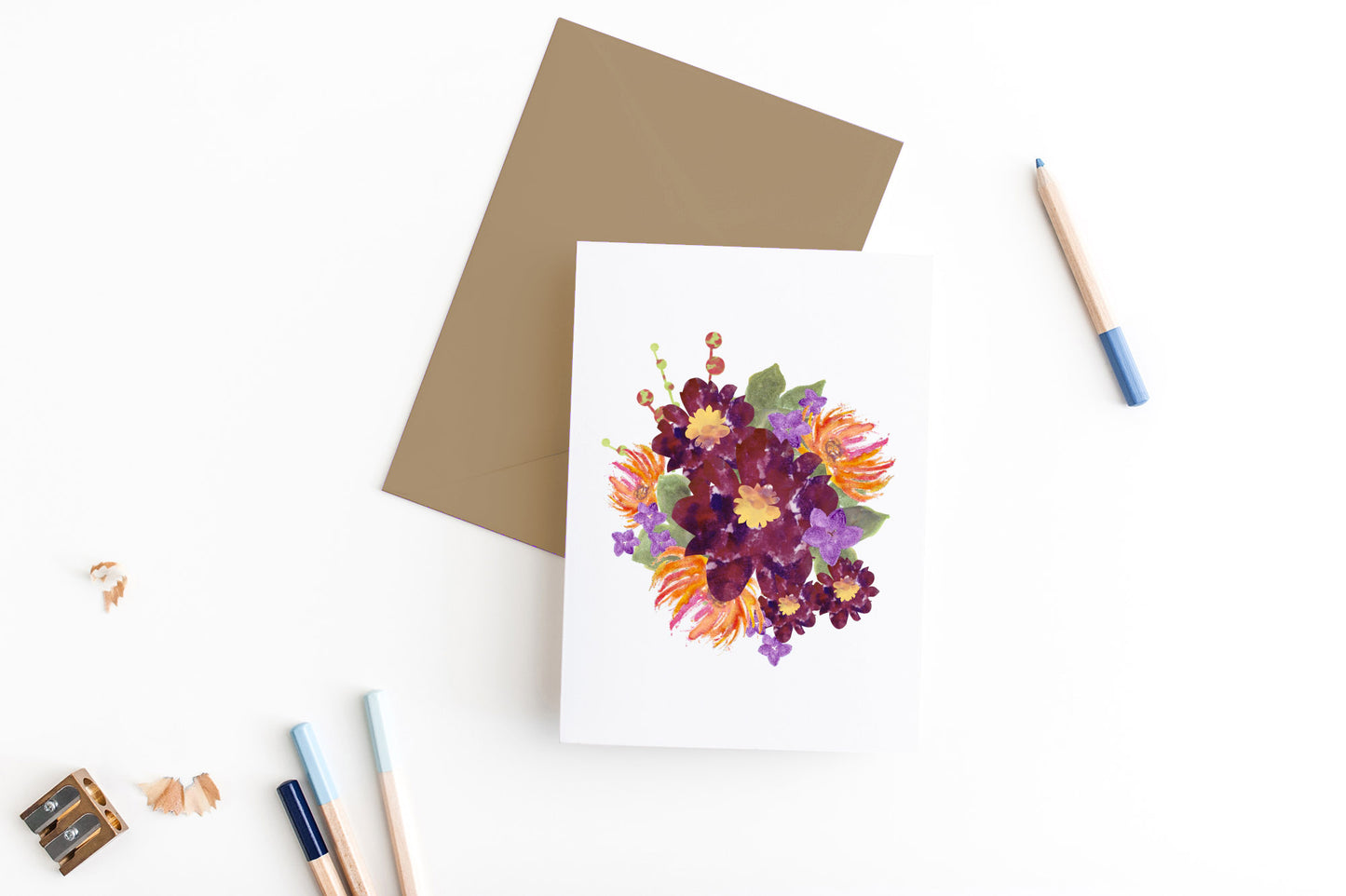 Greeting card, seen from above, with a kraft, square flap envelope, pencils, pencil sharpener and shavings on a white background.