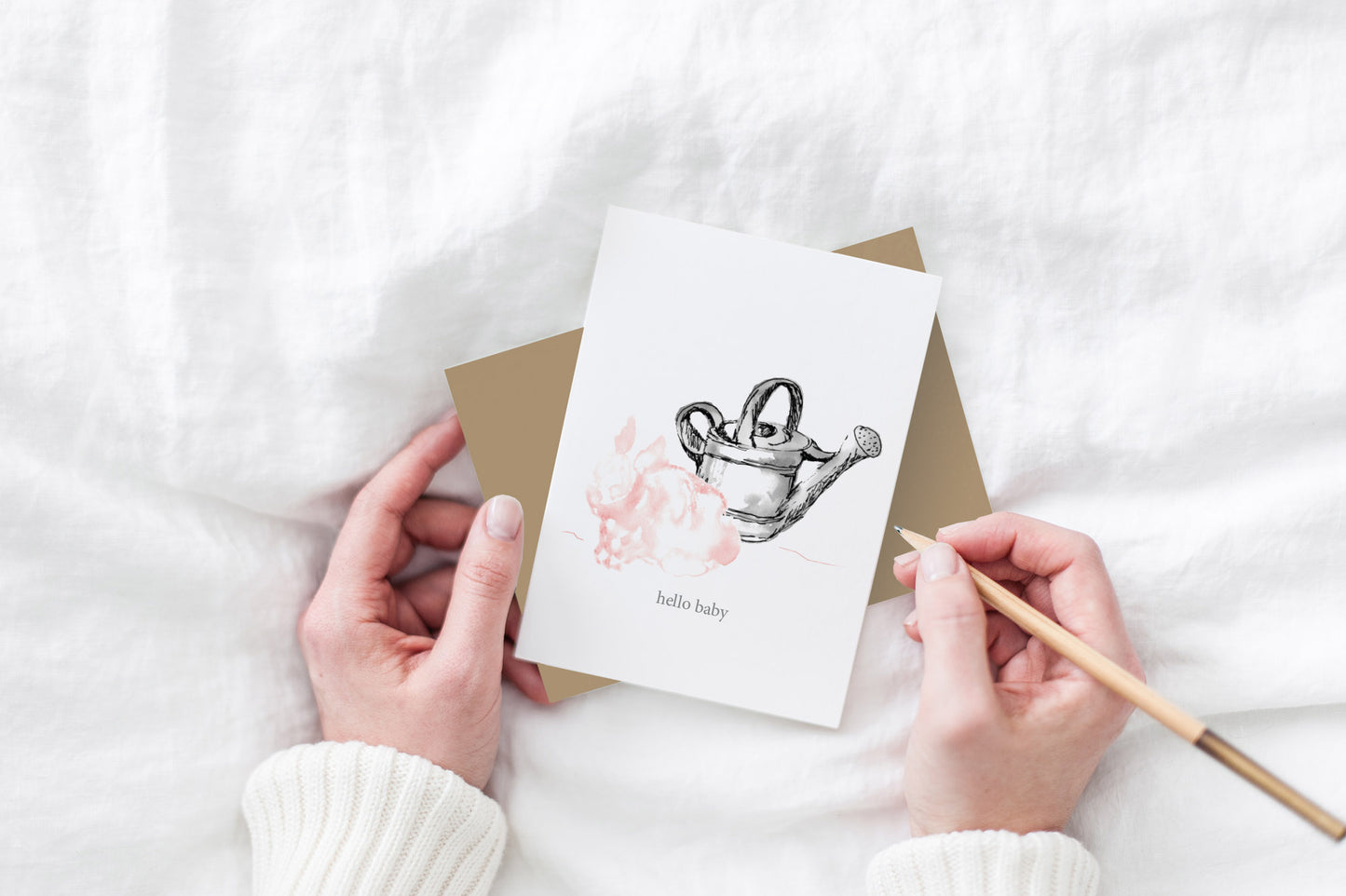Greeting card and kraft envelope, seen from above, held by two hands along with a pencil, ready to write, against a white comforter.