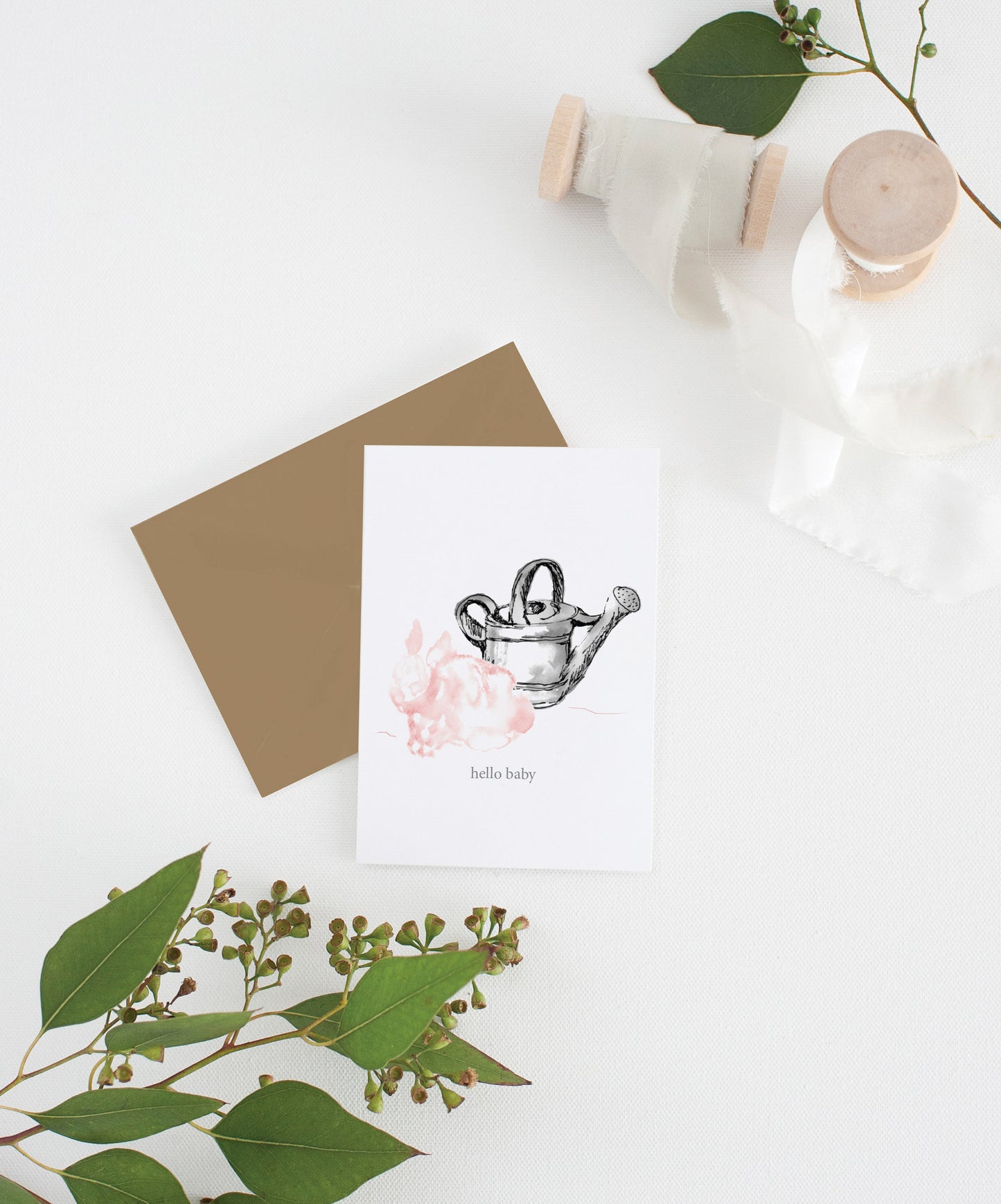 Greeting card with kraft envelope laying on a white surface with green leaves and cotton ribbon wound on wooden spools.