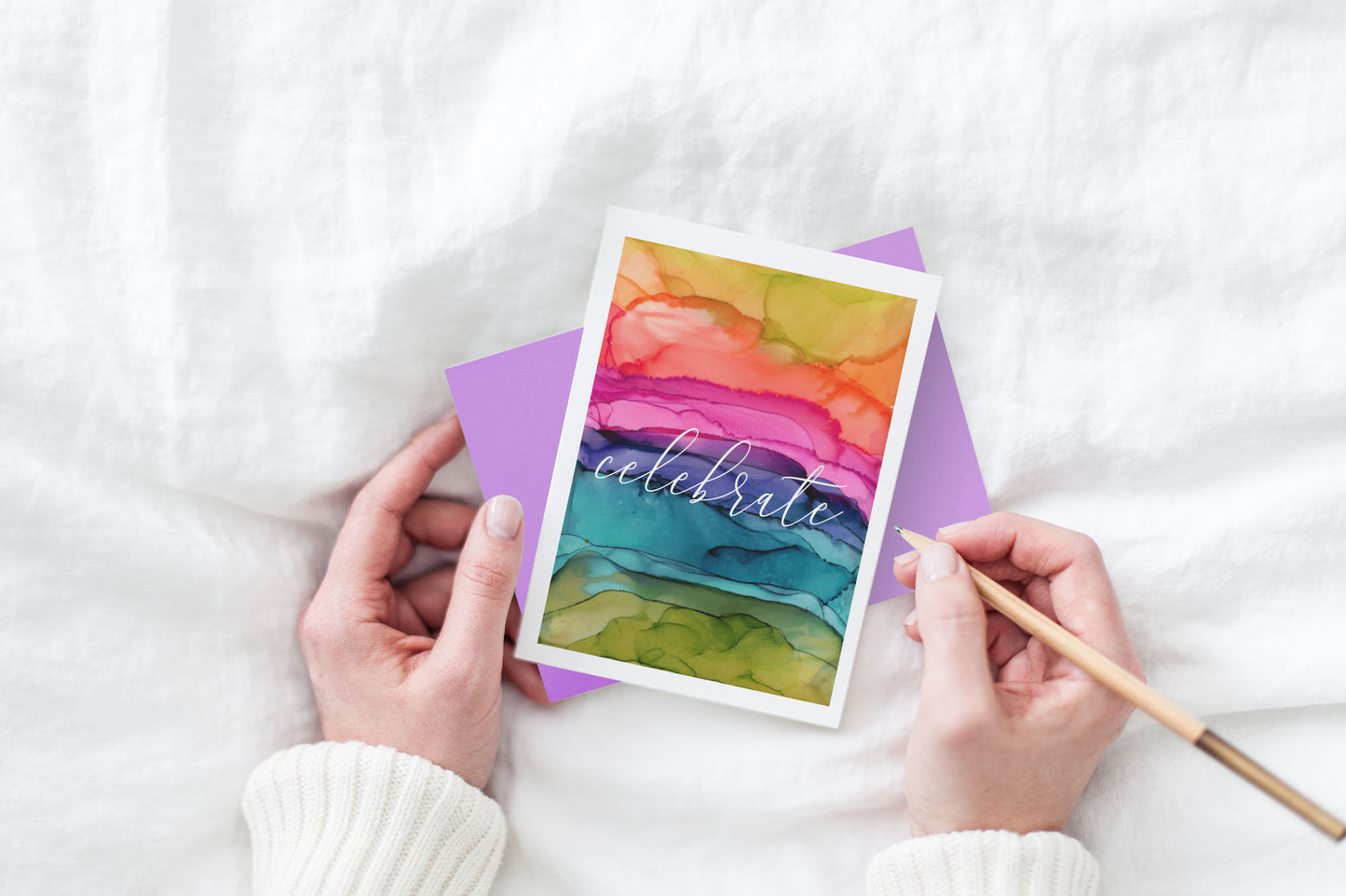 Greeting card and colorful envelope, seen from above, held by two hands along with a pencil, ready to write, against a white comforter.