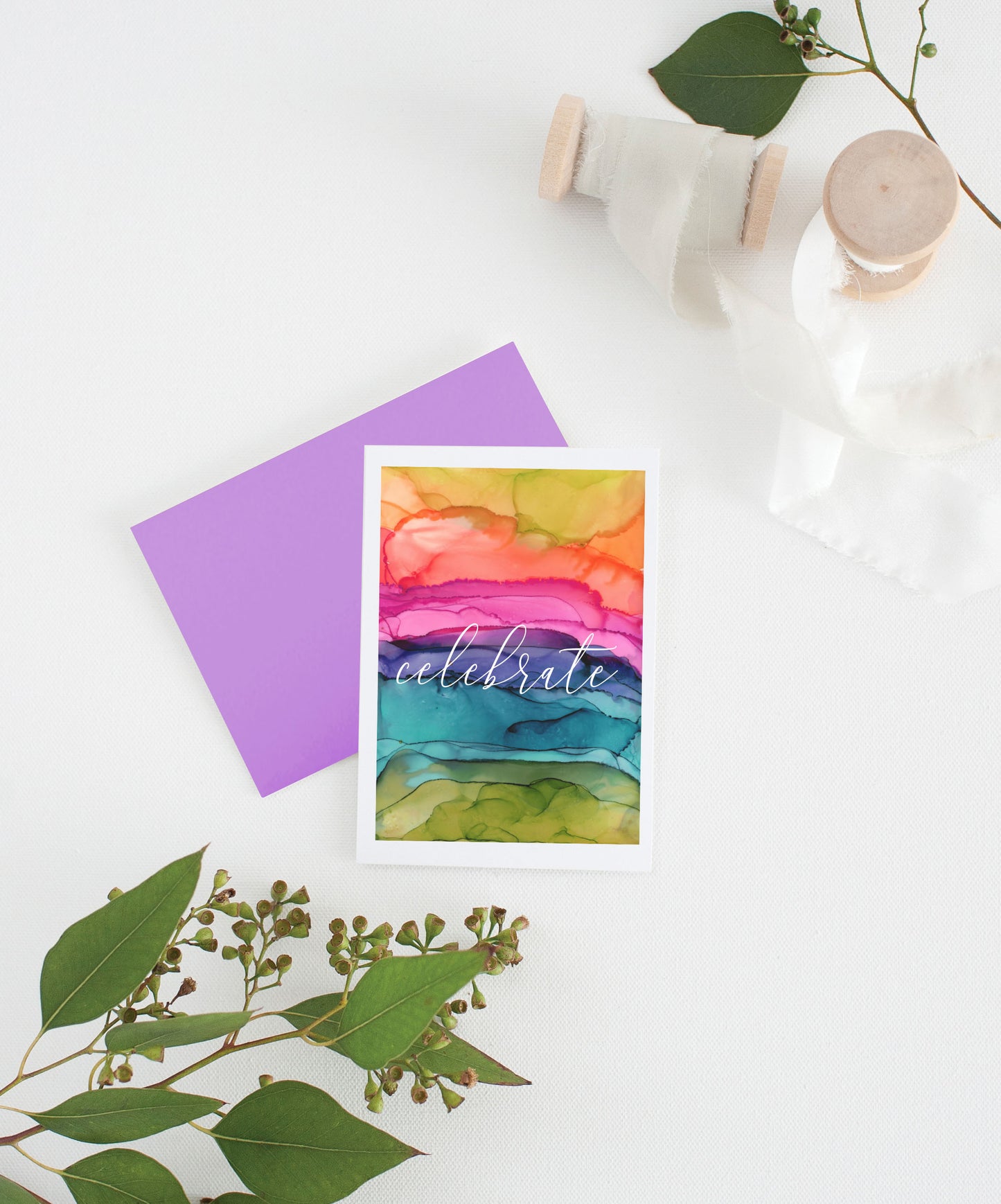 Greeting card with colorful envelope laying on a white surface with green leaves and cotton ribbon wound on wooden spools.