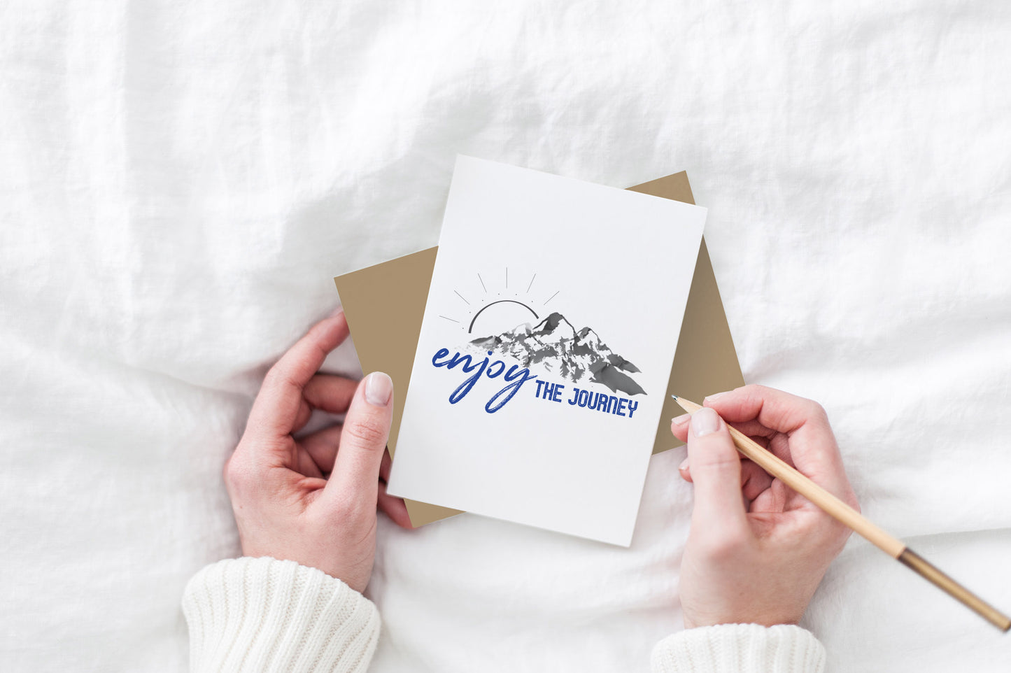 Greeting card and kraft envelope, seen from above, held by two hands along with a pencil, ready to write, against a white comforter.