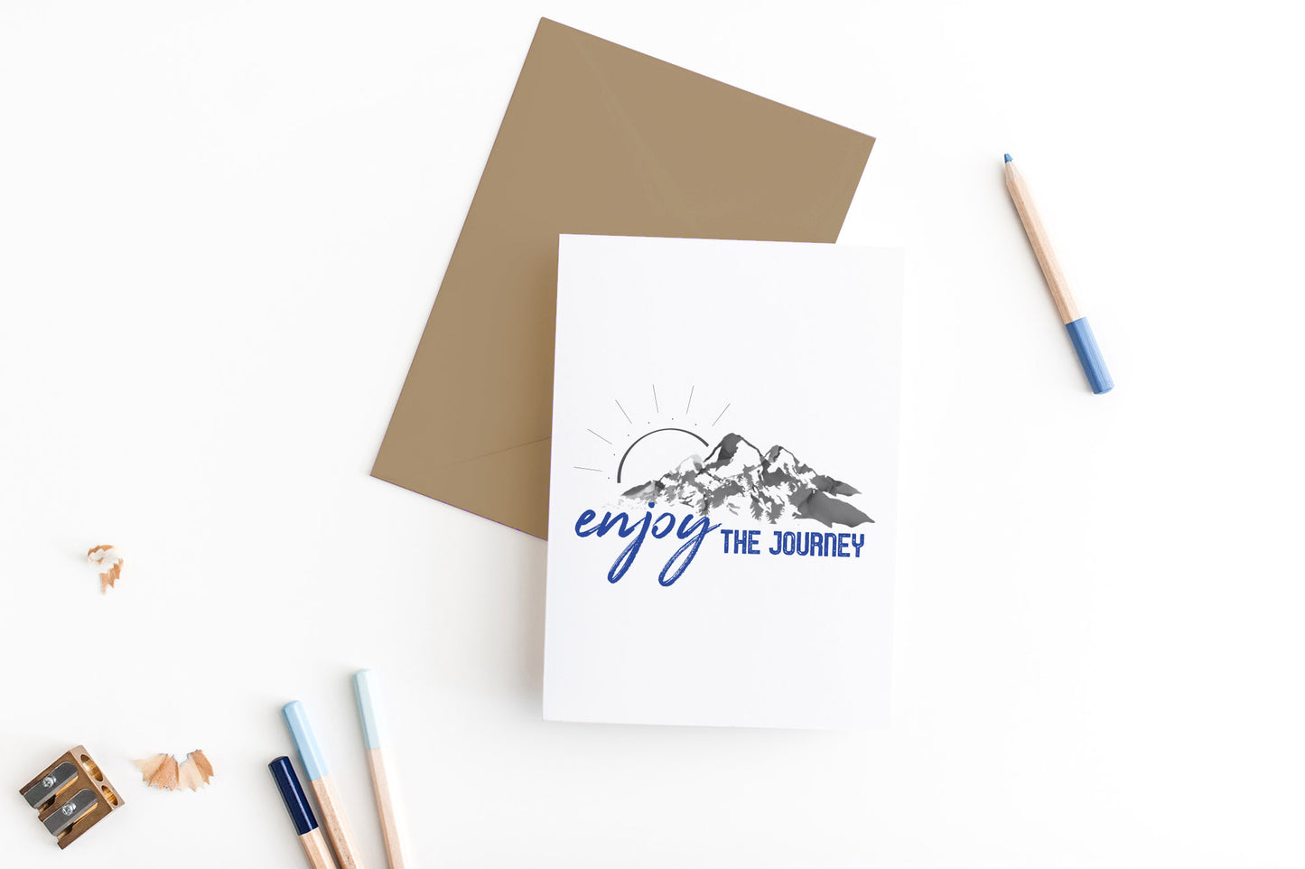 Greeting card, seen from above, with a kraft, square flap envelope, pencils, pencil sharpener and shavings on a white background.