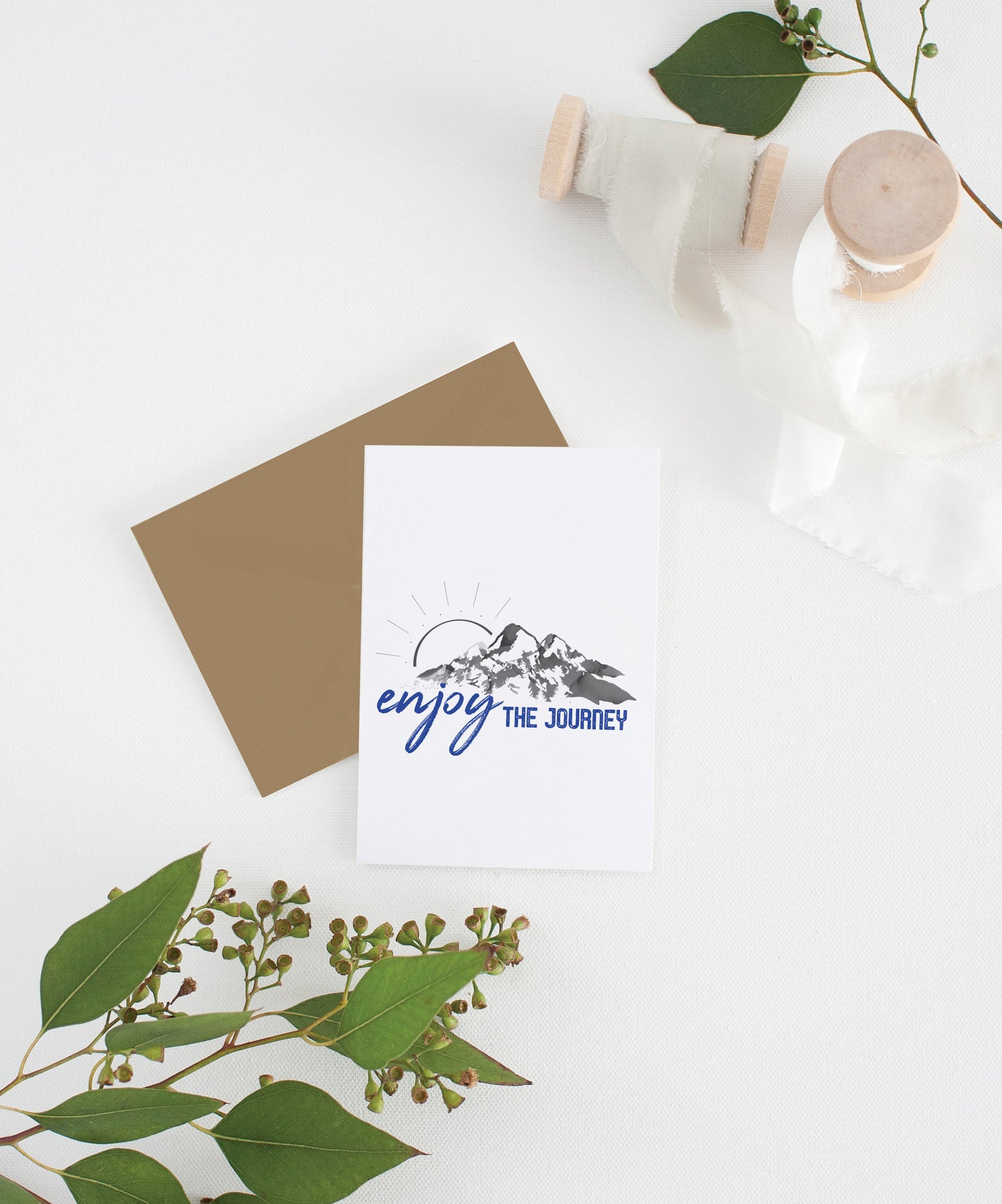 Greeting card with kraft envelope laying on a white surface with green leaves and cotton ribbon wound on wooden spools.