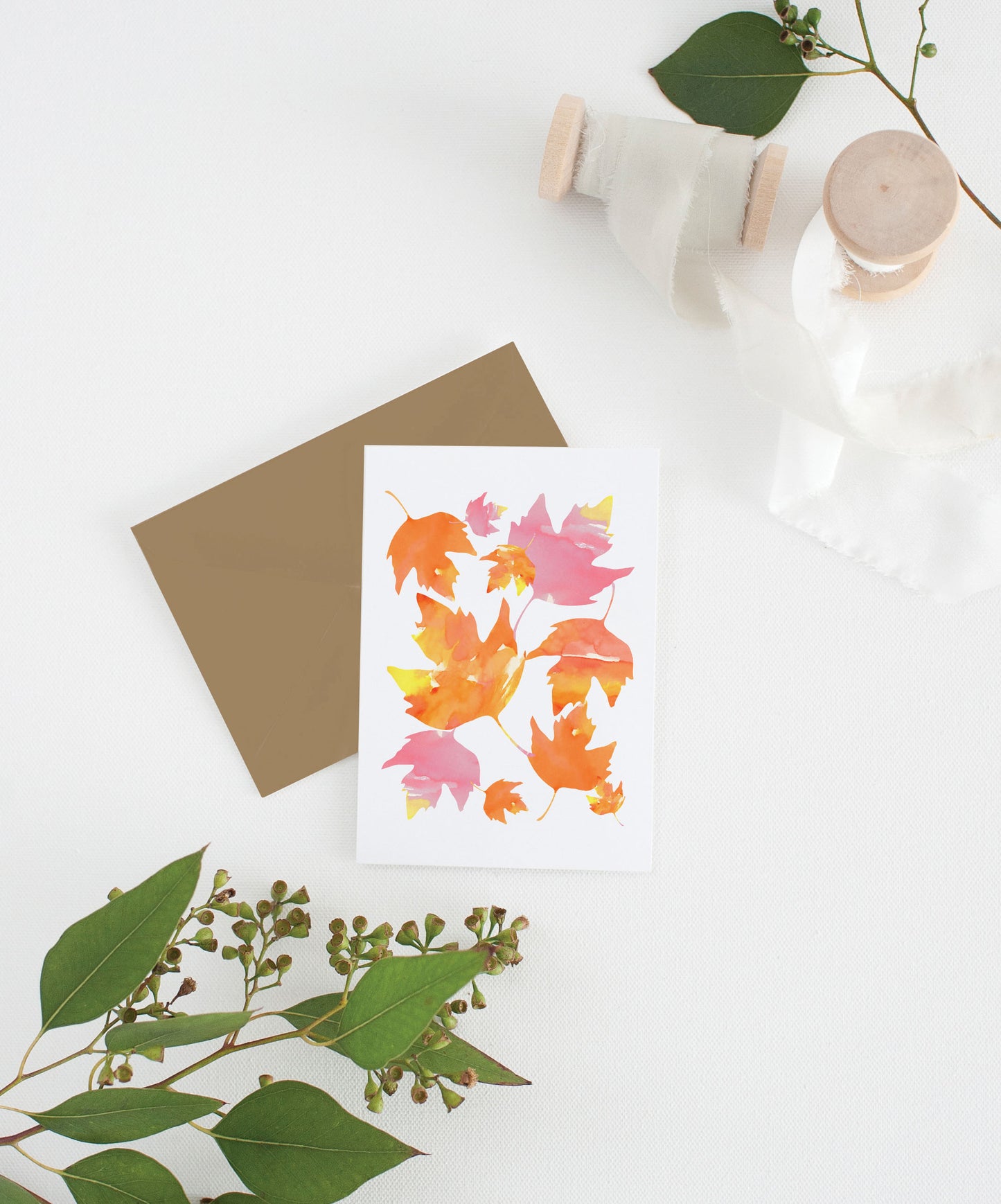 Greeting card with kraft envelope laying on a white surface with green leaves and cotton ribbon wound on wooden spools.