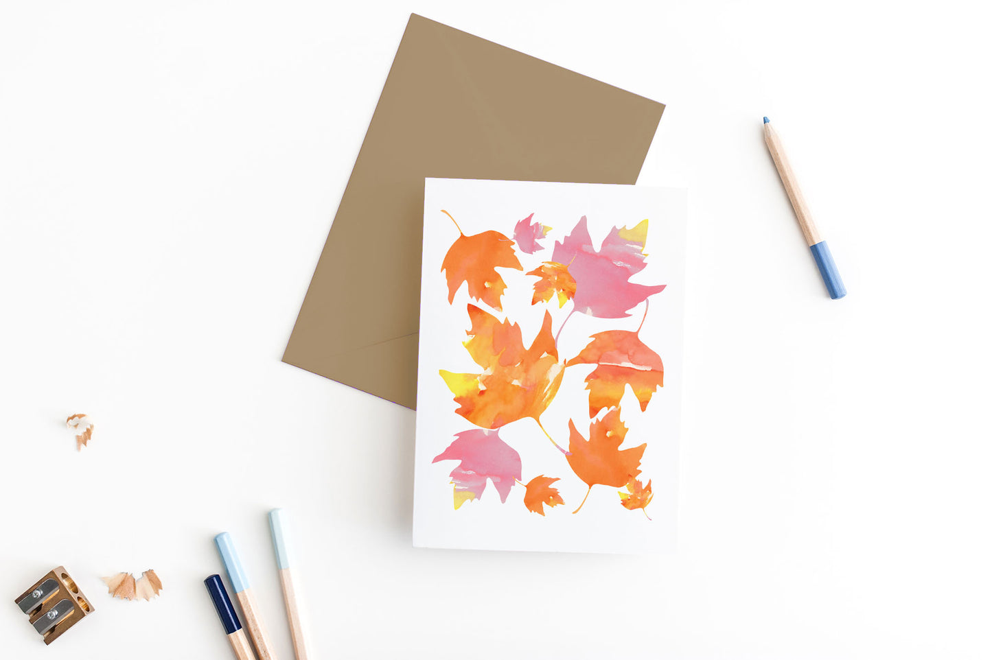Greeting card, seen from above, with a kraft, square flap envelope, pencils, pencil sharpener and shavings on a white background.