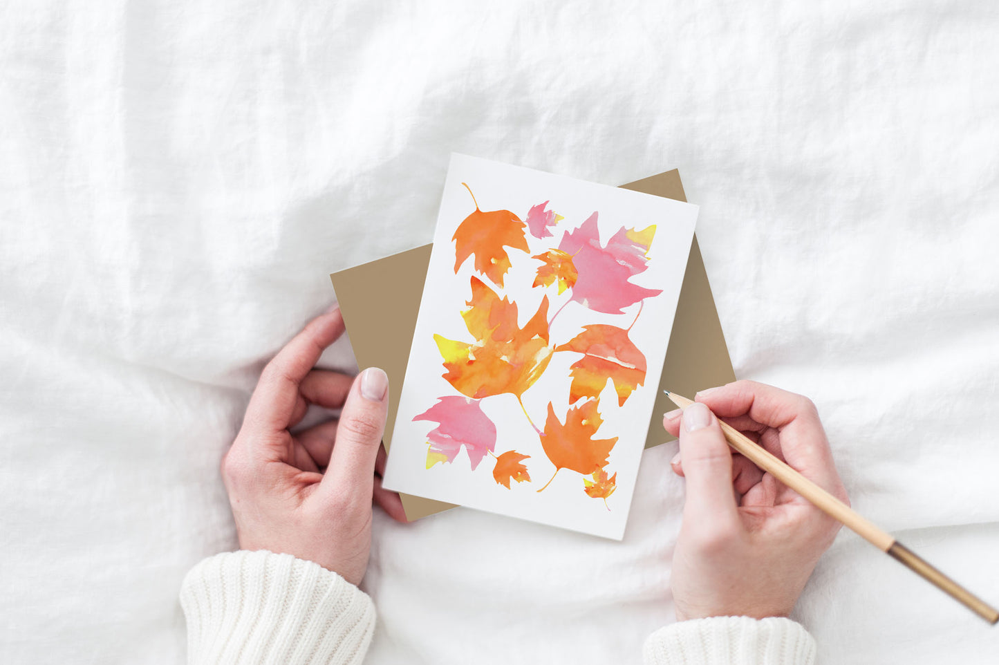 Greeting card and kraft envelope, seen from above, held by two hands along with a pencil, ready to write, against a white comforter.