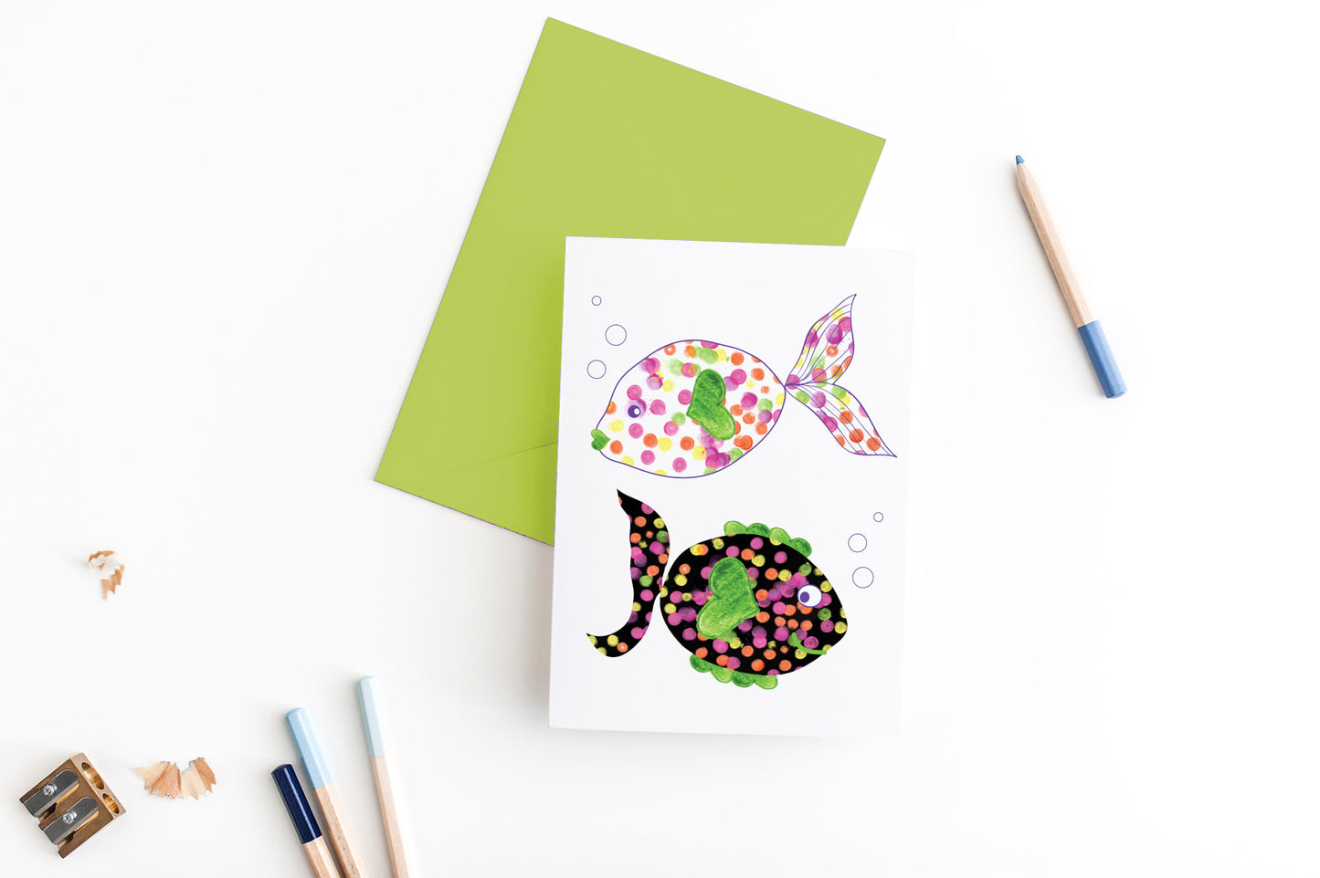Greeting card, seen from above, with a colored, square flap envelope, pencils, pencil sharpener and shavings on a white background.