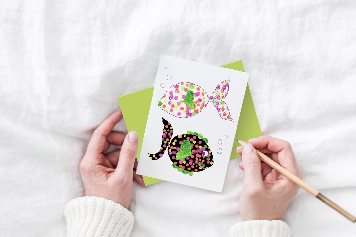 Greeting card and colorful envelope, seen from above, held by two hands along with a pencil, ready to write, against a white comforter.