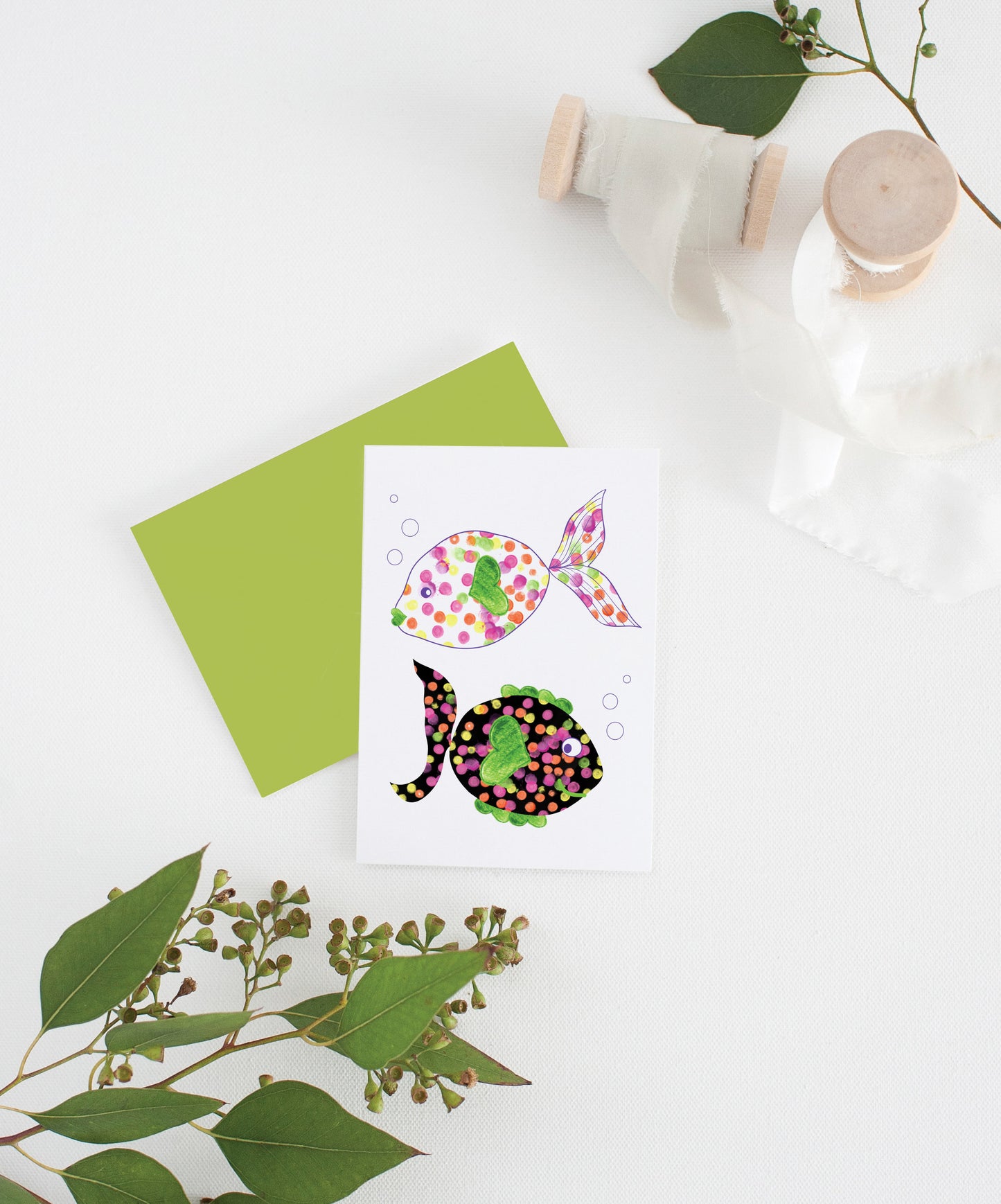 Greeting card with colorful envelope laying on a white surface with green leaves and cotton ribbon wound on wooden spools.