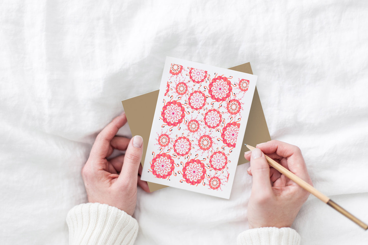 Greeting card and kraft envelope, seen from above, held by two hands along with a pencil, ready to write, against a white comforter.