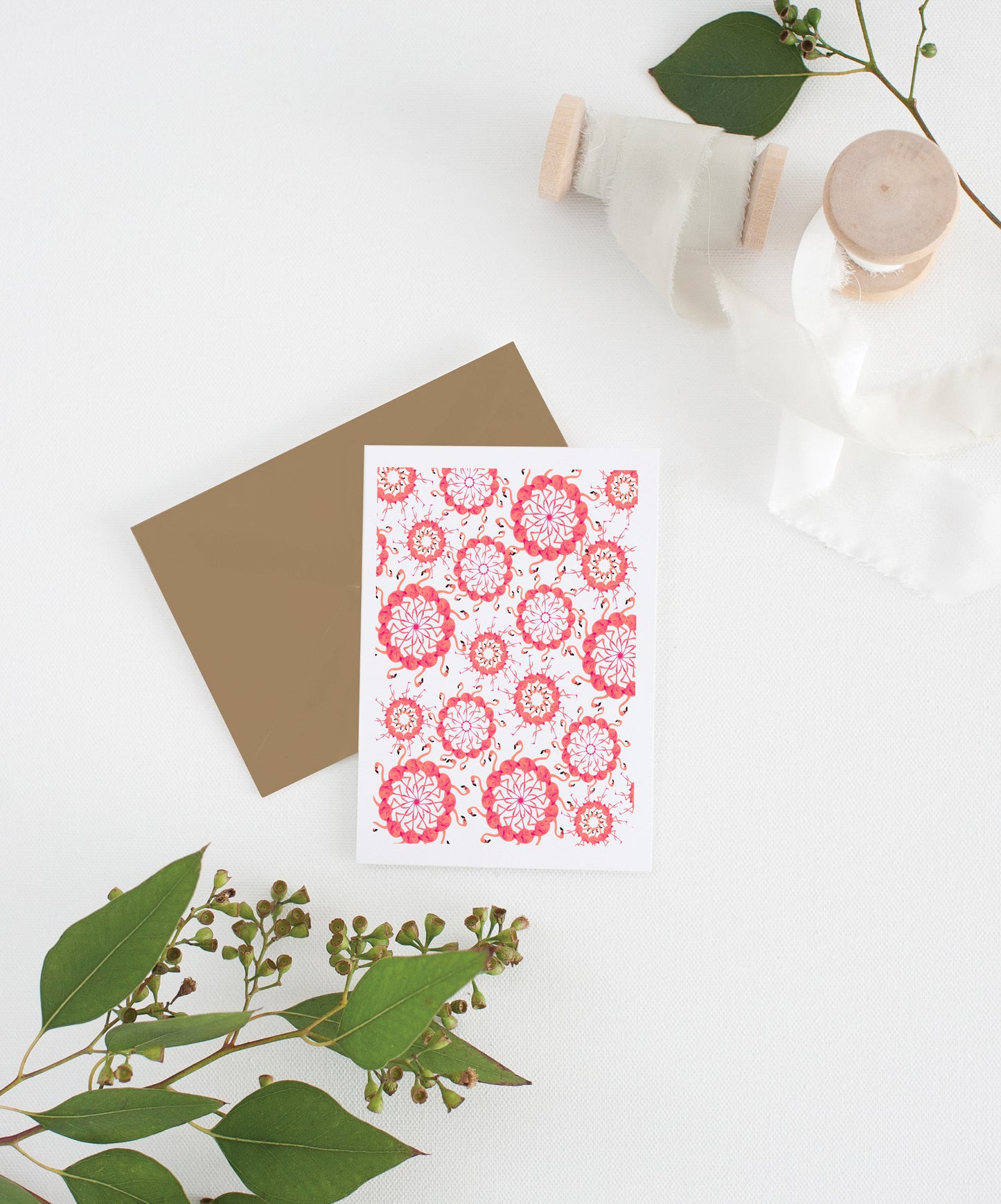 Greeting card with kraft envelope laying on a white surface with green leaves and cotton ribbon wound on wooden spools.