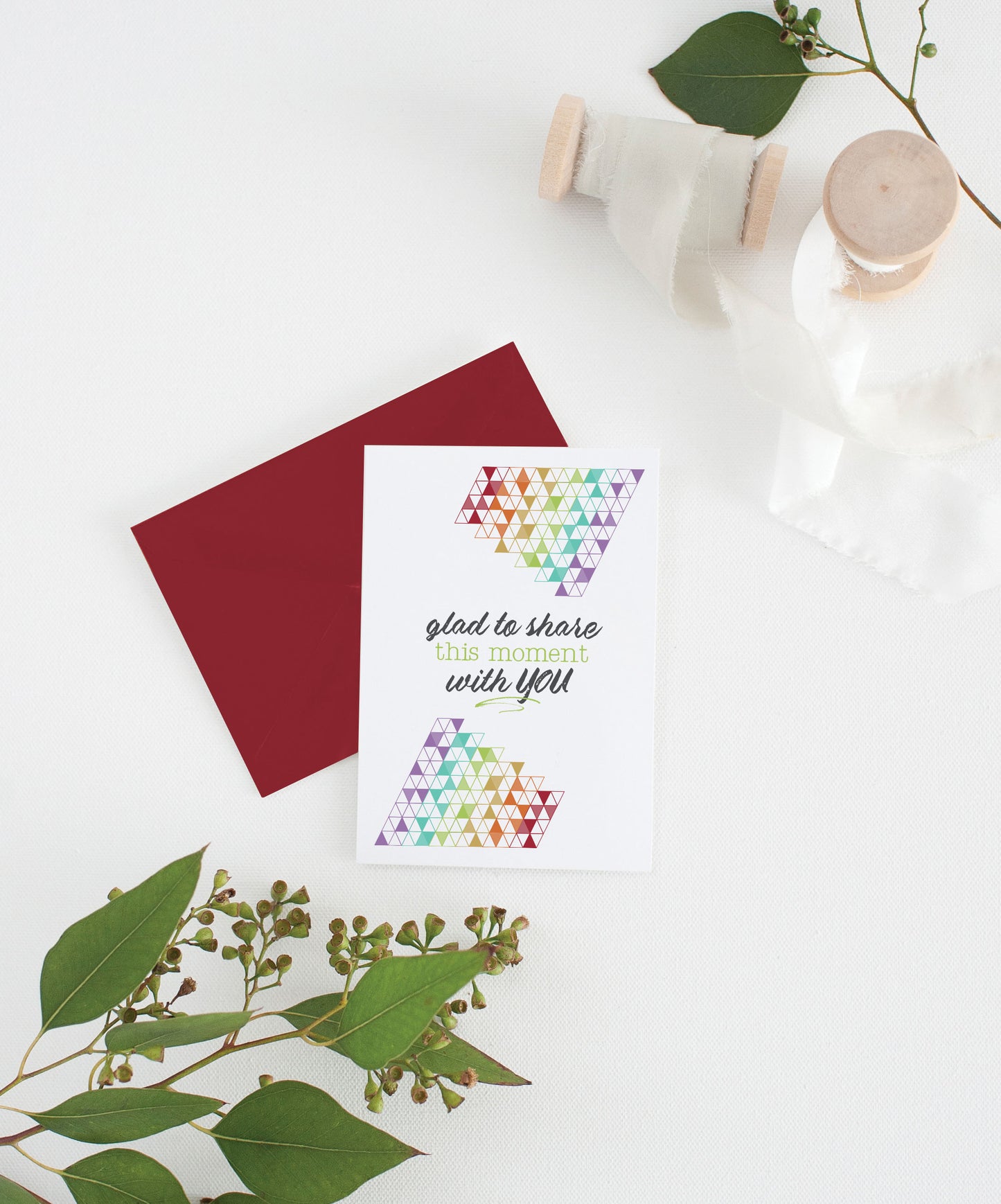 Greeting card with colorful envelope laying on a white surface with green leaves and cotton ribbon wound on wooden spools.