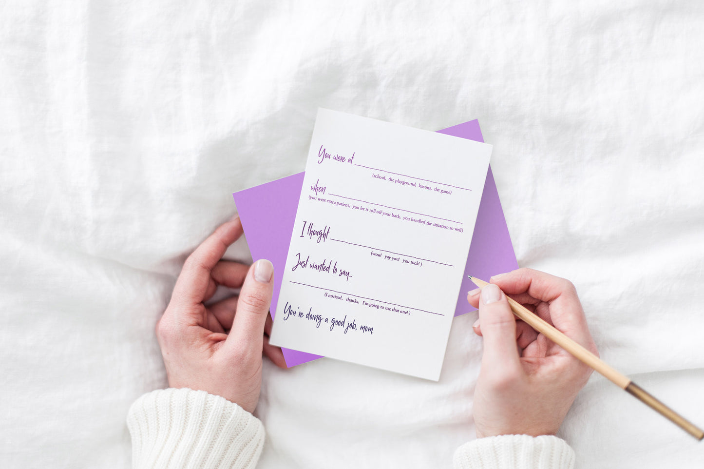 Greeting card and colorful envelope, seen from above, held by two hands along with a pencil, ready to write, against a white comforter.